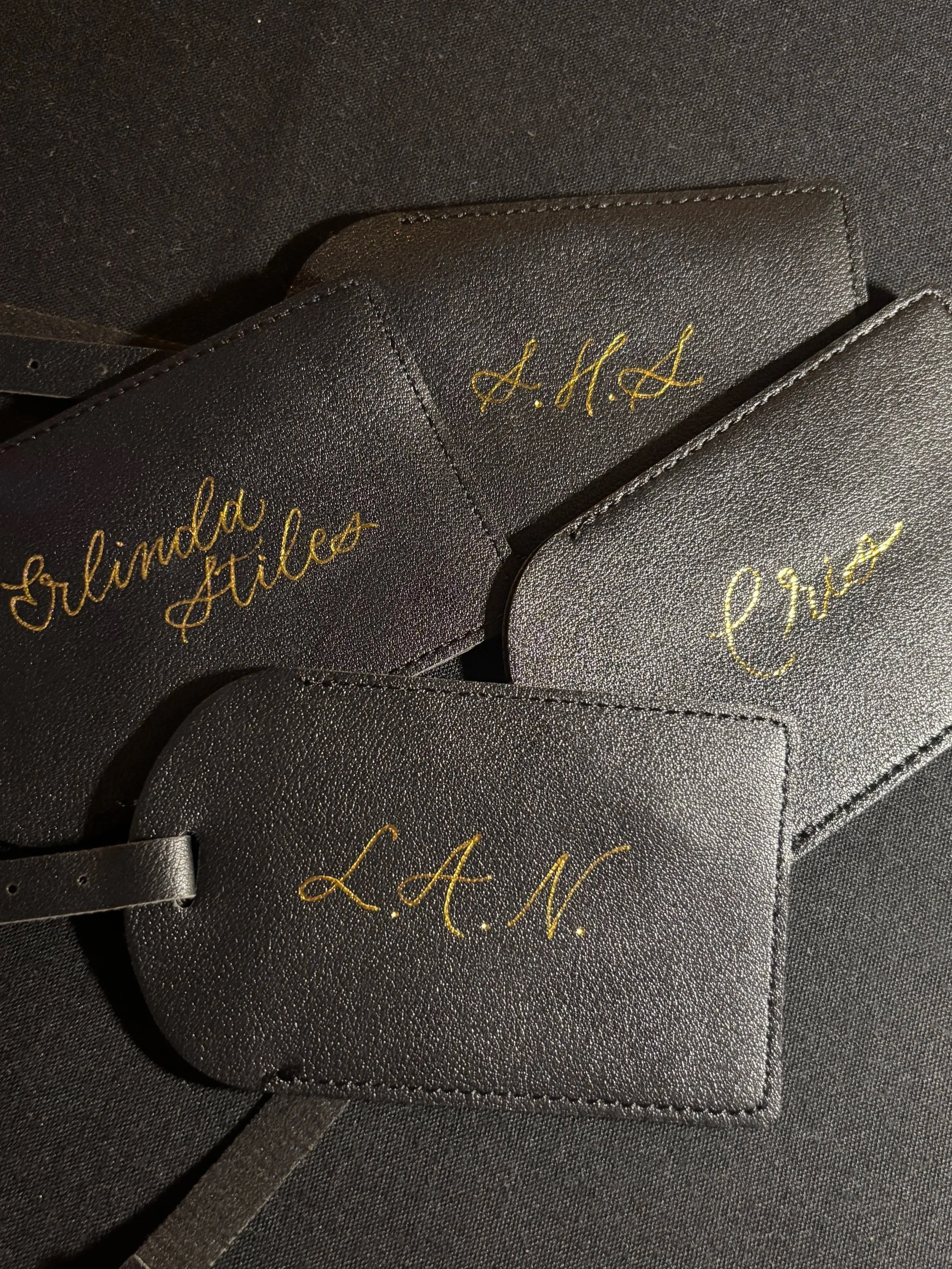 Black leather luggage tags with gold embroidered initials and names: 'L. A. N.', 'E. R. A. L.', and 'Glinda Miles'.