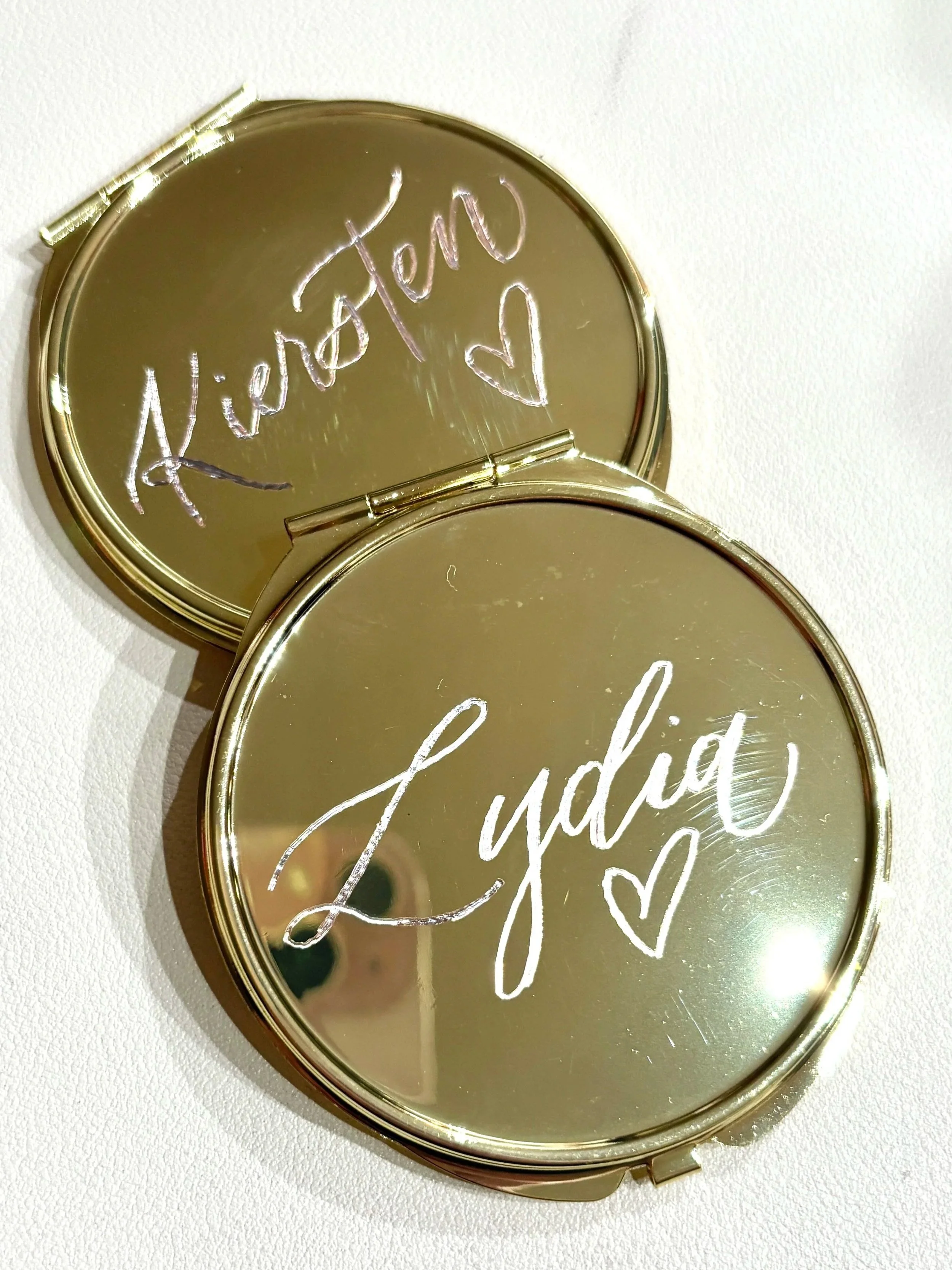 A round compact mirror with a gold frame, with hand-engraved text and hearts on it, placed on a white surface.