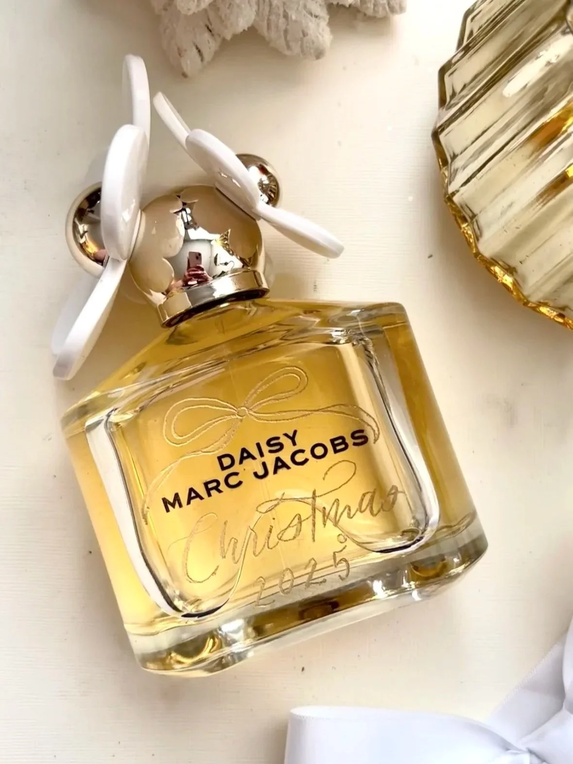 A Marc Jacobs Daisy perfume bottle with a silver cap and a white bow attached, placed on a white surface.