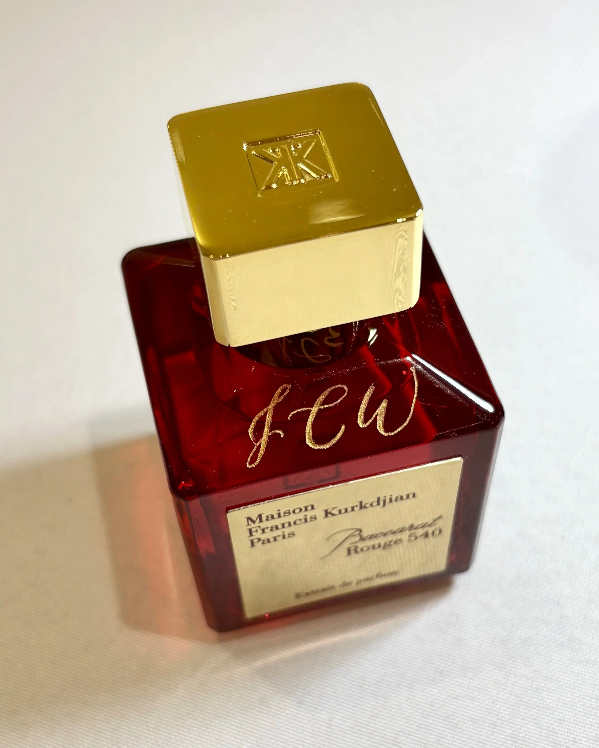 A red bottle of Maison Francis Kurkdjian Beijing Rouge 540 perfume with a gold and cream cap, on a white surface with the letters J,C,W engraved on the top.
