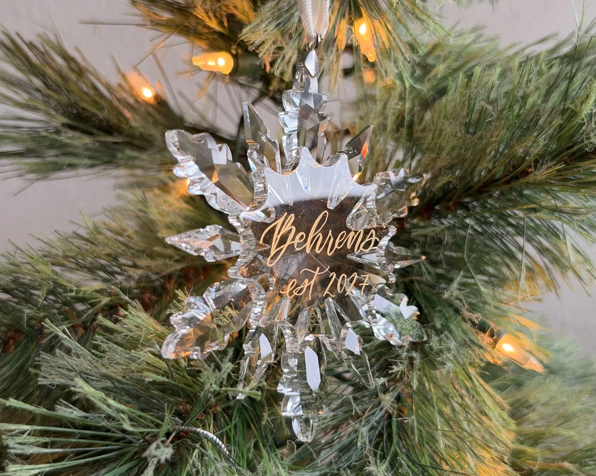 A crystal snowflake ornament with the words "Behrens est. 2027" in gold lettering, in front of an evergreen christmas tree.