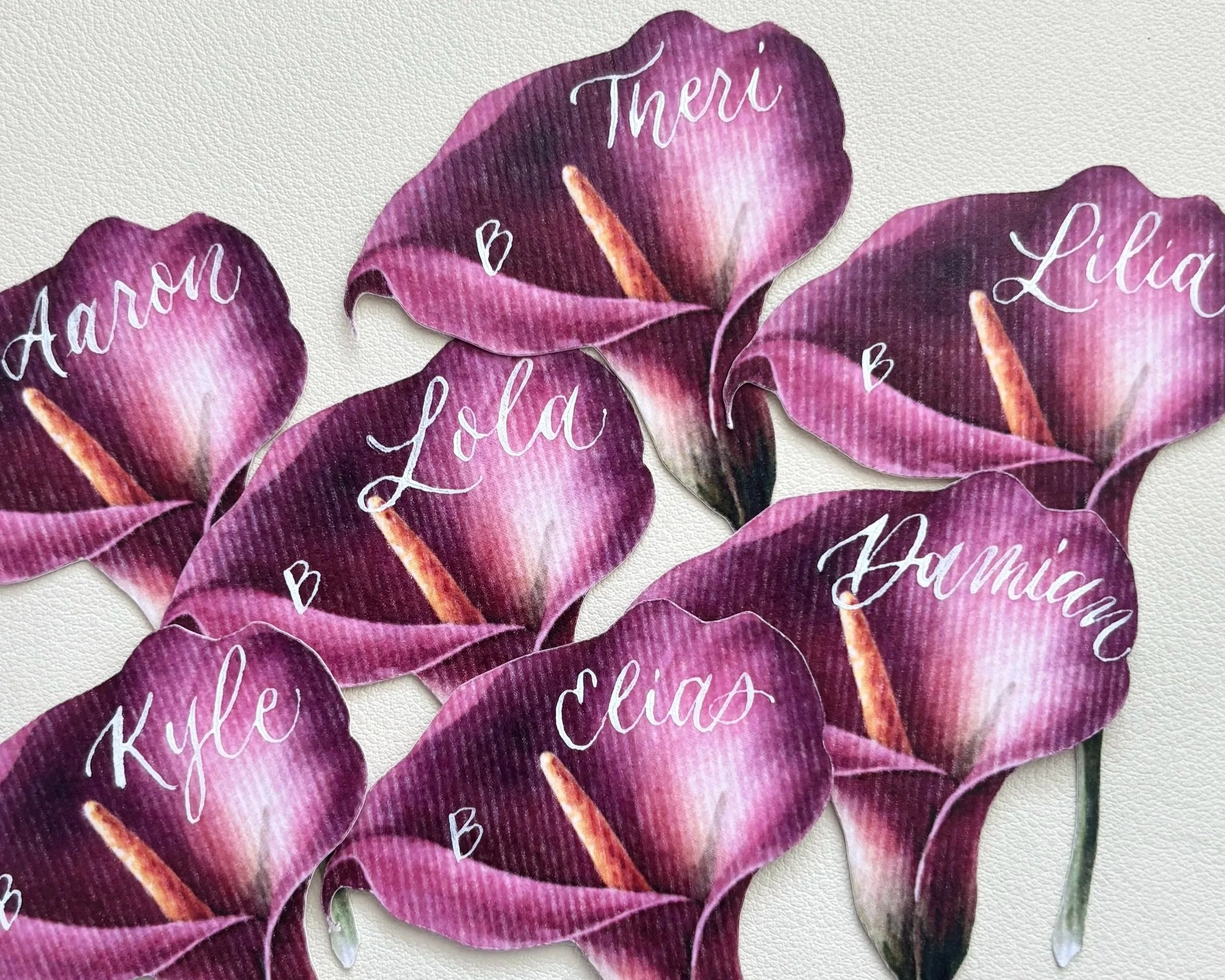 A group of wedding place cards on paper shaped like calla lilies with names written in calligraphy in white ink.