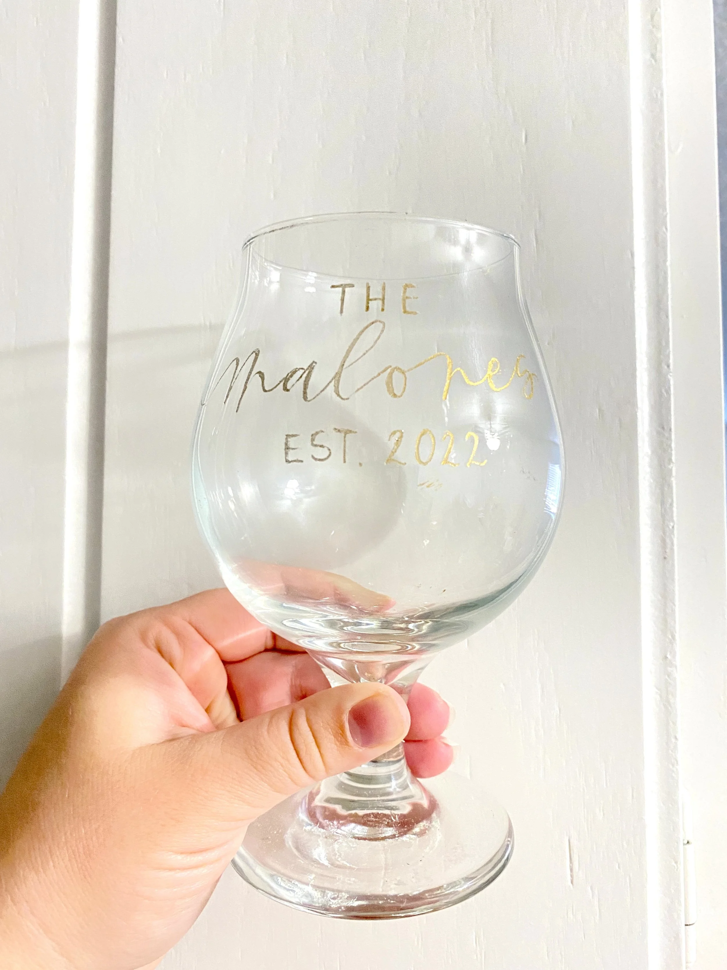 Hand holding a clear glass with gold writing that says 'The Malones, Est. 2022' against a white wall background.
