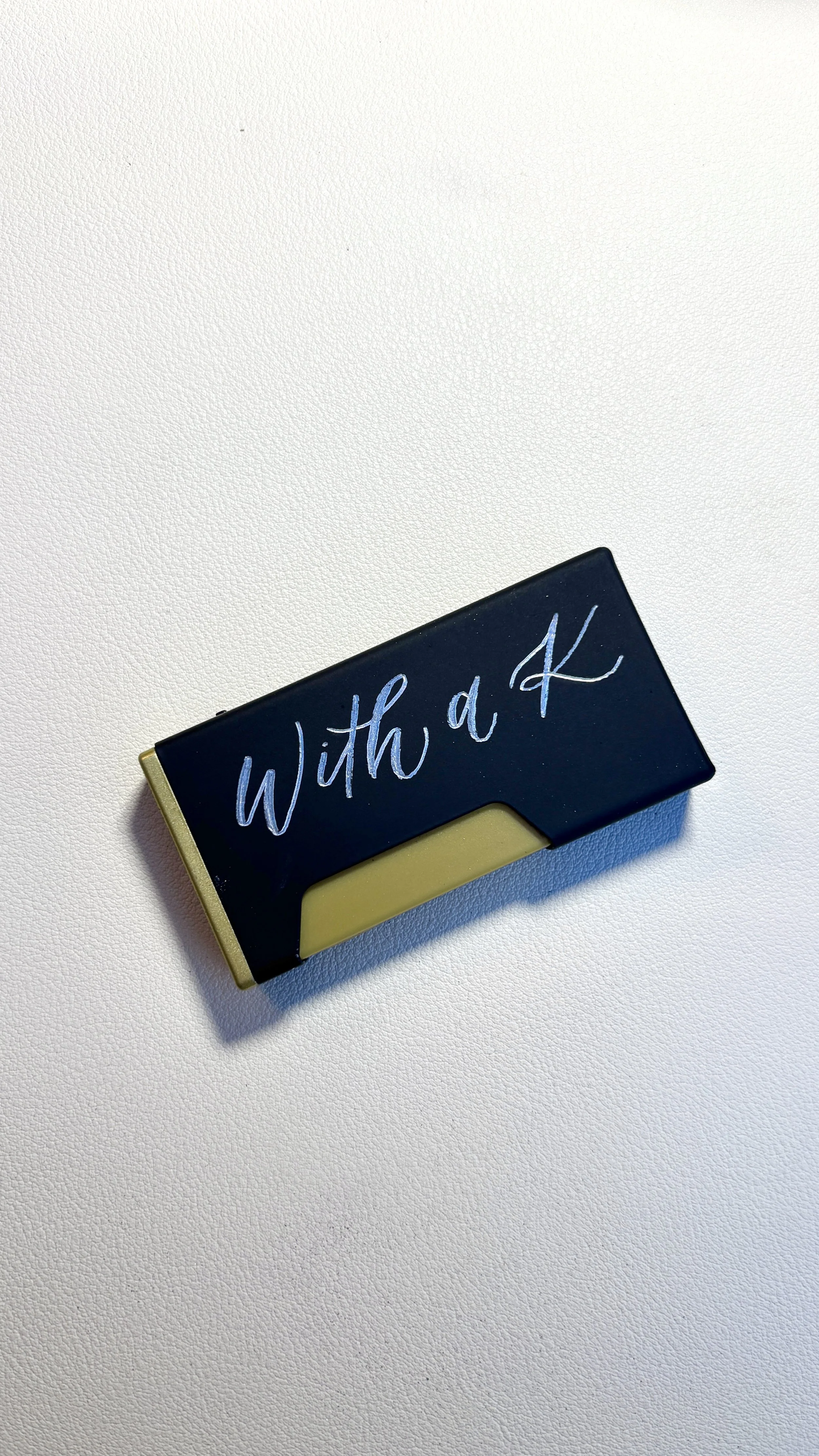 Close-up of a black and gold lighter with the words 'With a K' written in cursive on the side, placed on a plain white surface.