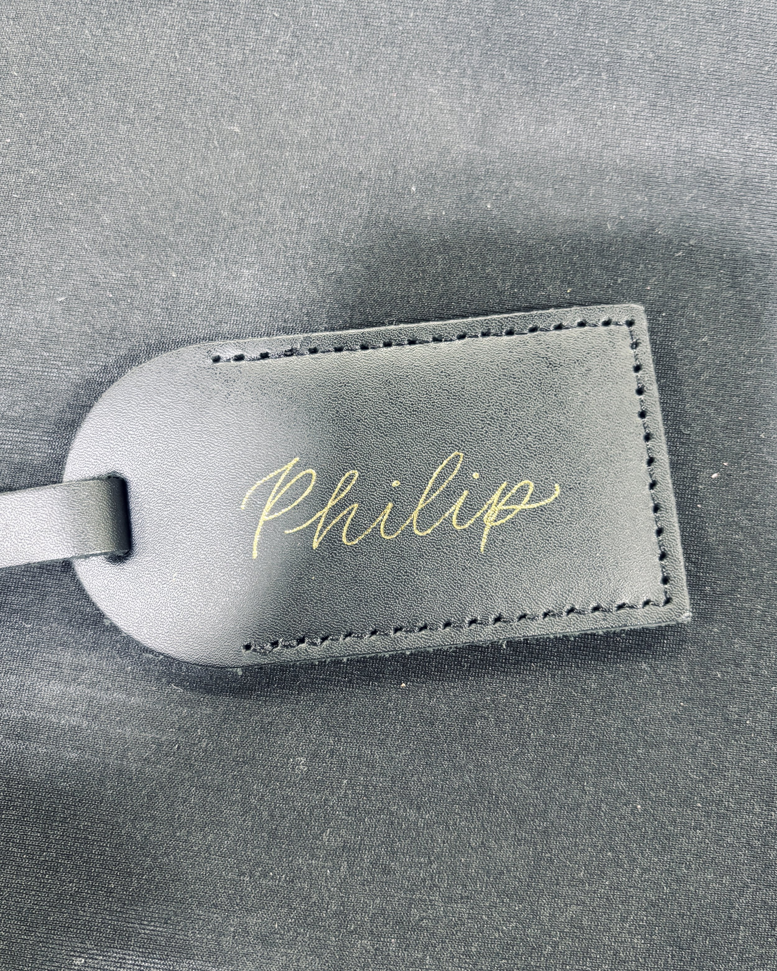 Luggage tag personalization in Denver, CO Close-up of a black leather luggage tag with the name "Philip" written in gold cursive, attached to a black surface.