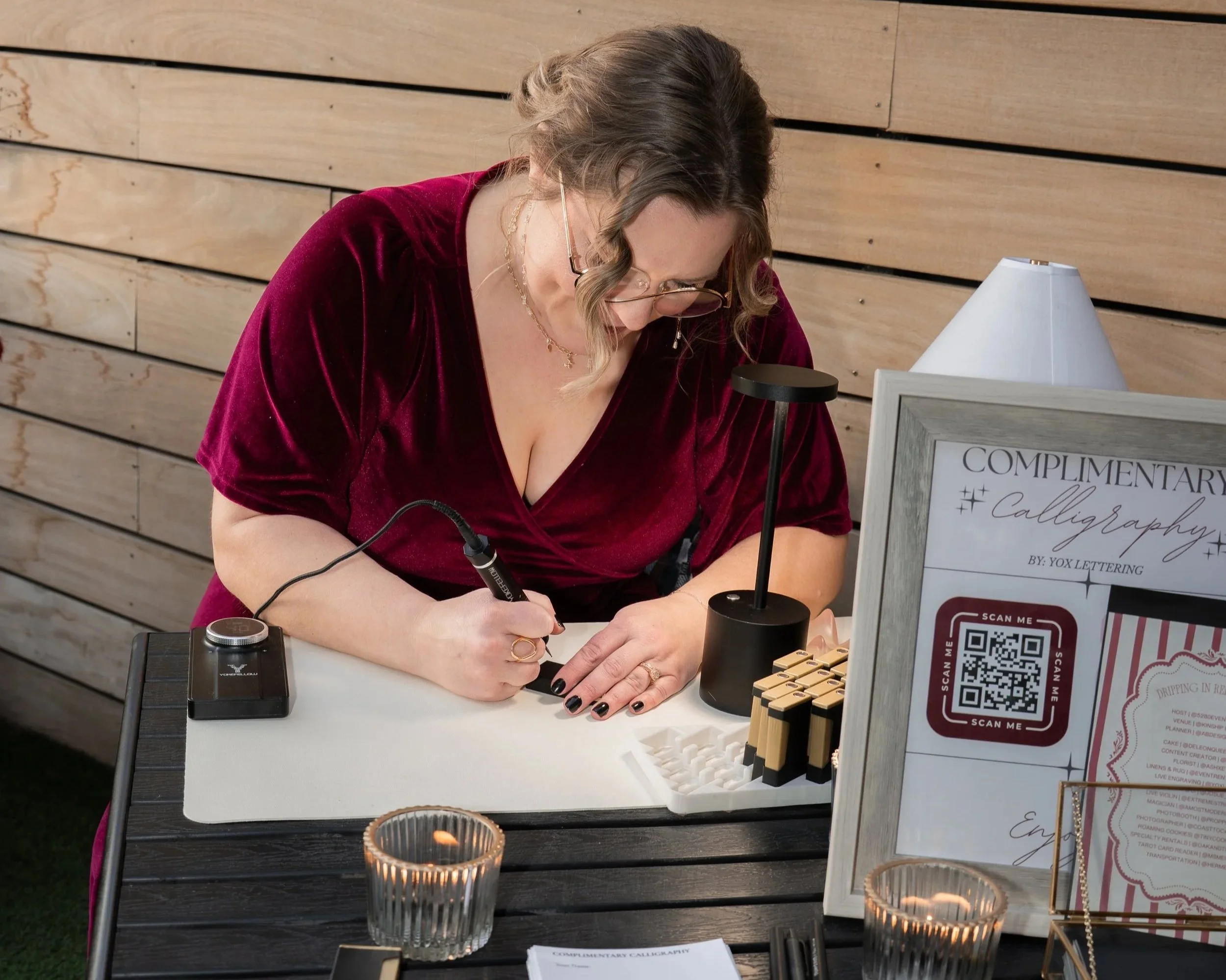 Woman in a burgundy dress practicing calligraphy at a table outside.