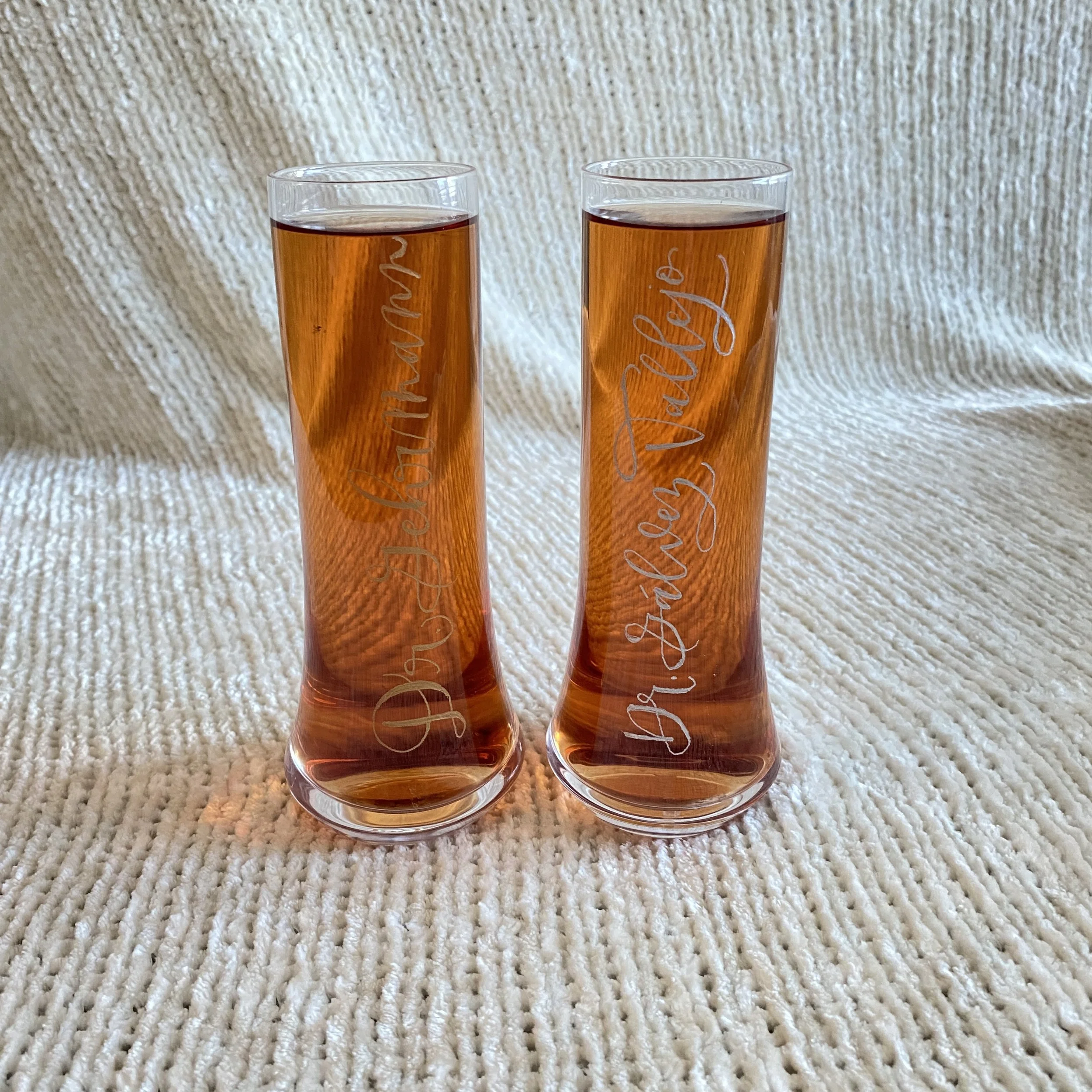 Two tall, narrow glass vases filled with orange liquid, placed on a beige textured fabric background.
