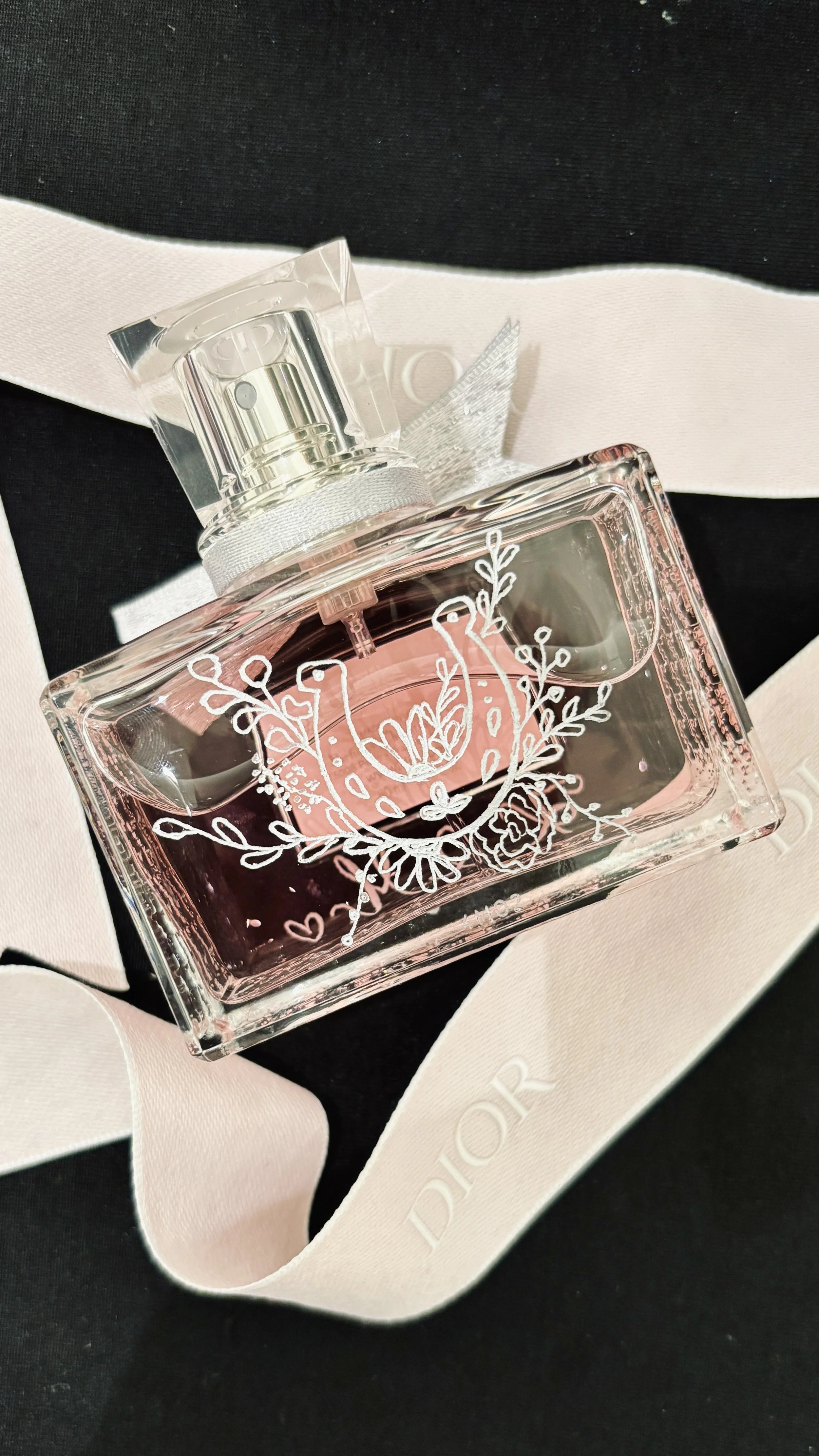 A DIOR perfume bottle with pink liquid, decorated with white floral designs, placed on a white ribbon with the word "DIOR" on it, against a black background.