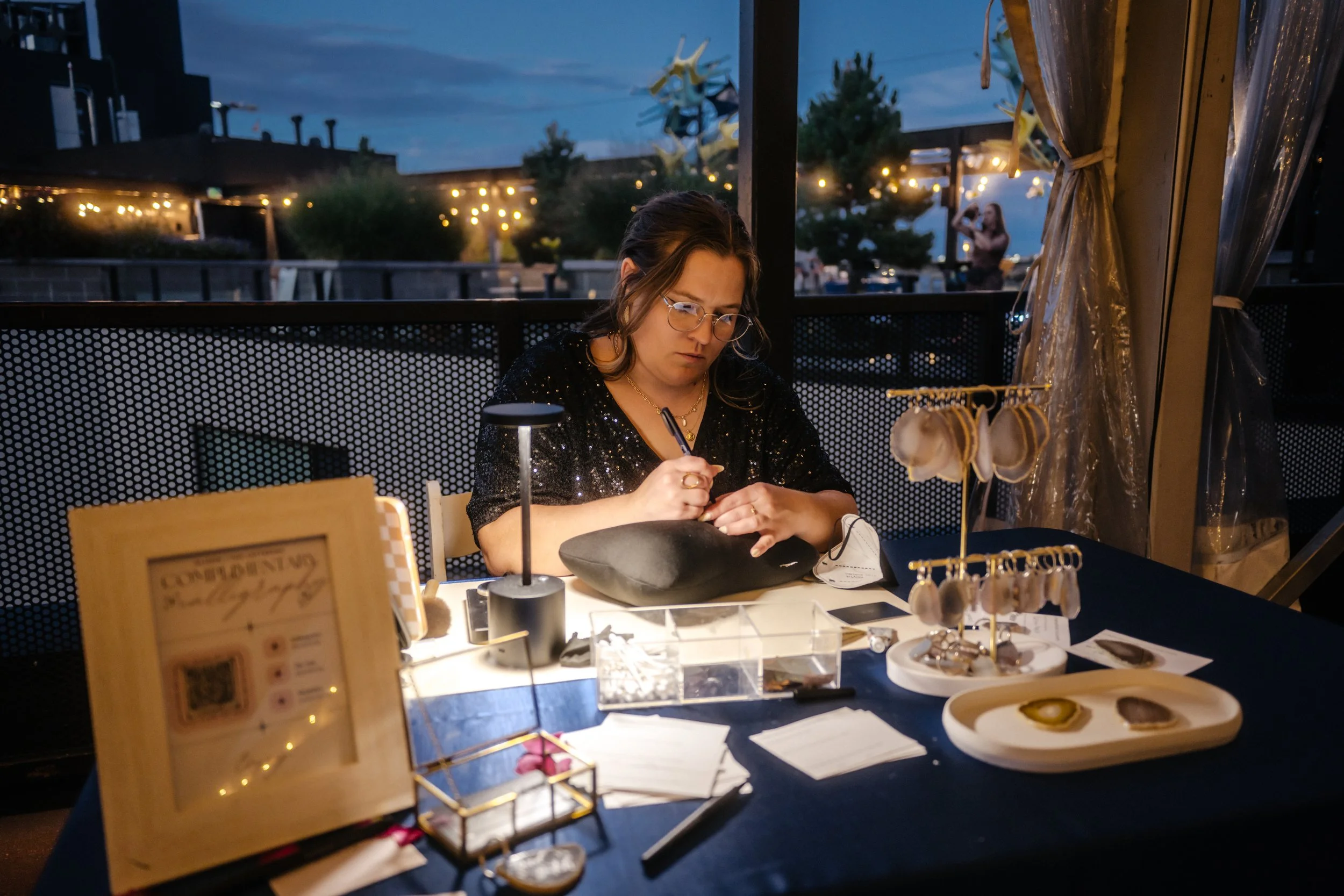 A woman dressed in all black is hand engraving an agate keychain for a wedding at a rooftop in Denver, CO.