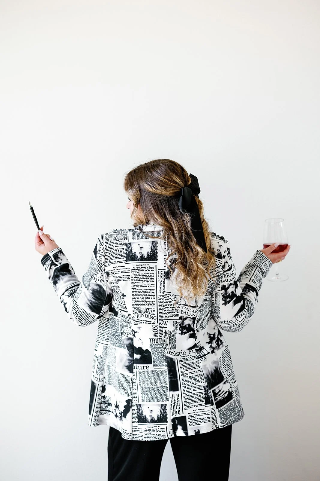 Alison is wearing a newspaper print jacket and holding a glass of red wine in one hand and a calligraph pen in the other. She's standing in front of a white wall.