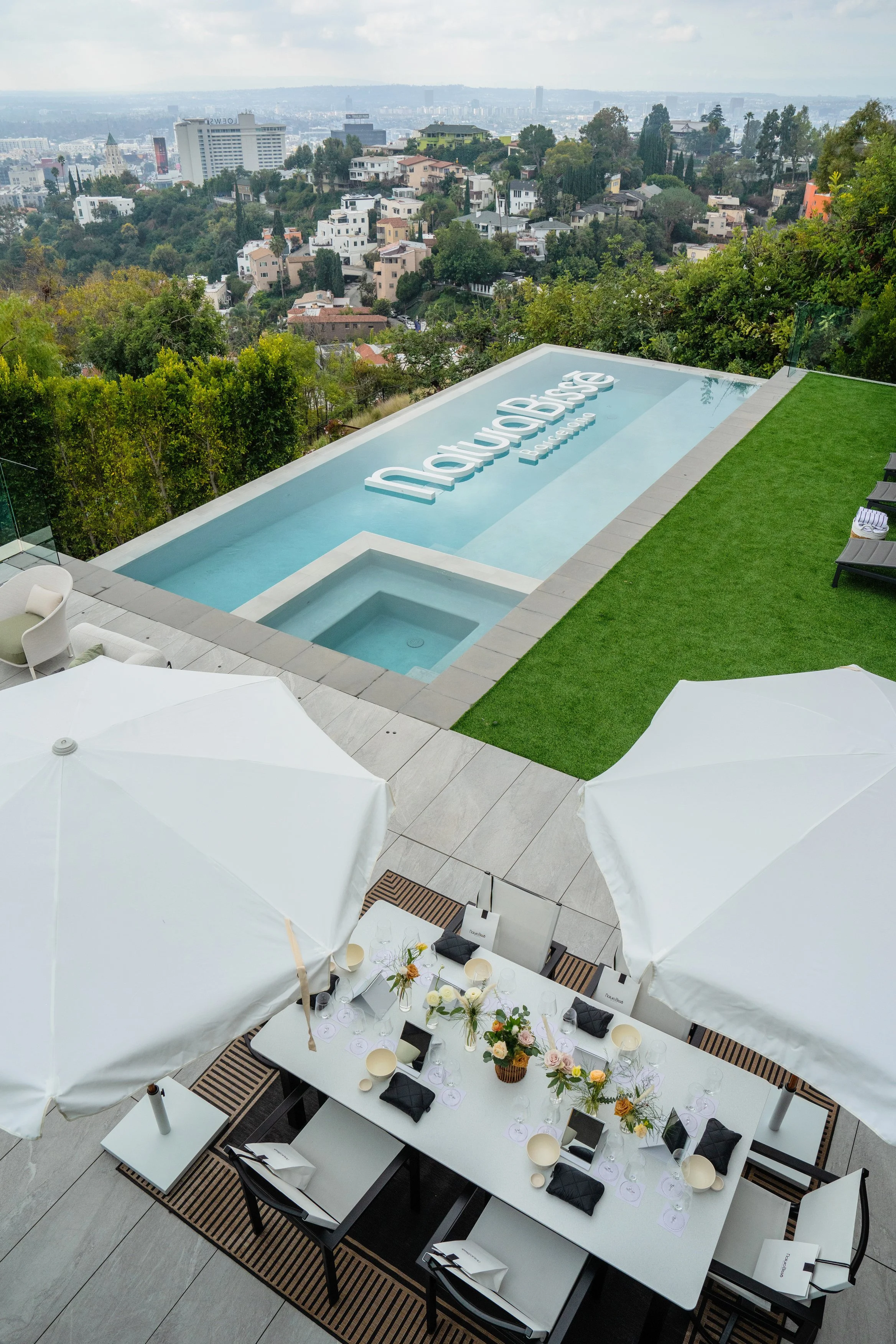 Overlooking a city from a patio featuring a rectangular pool with the branding 'Love Bisse' in the water. The patio has a dining area with tables, chairs, umbrellas, and floral arrangements, with lush greenery and city skyline in the background.