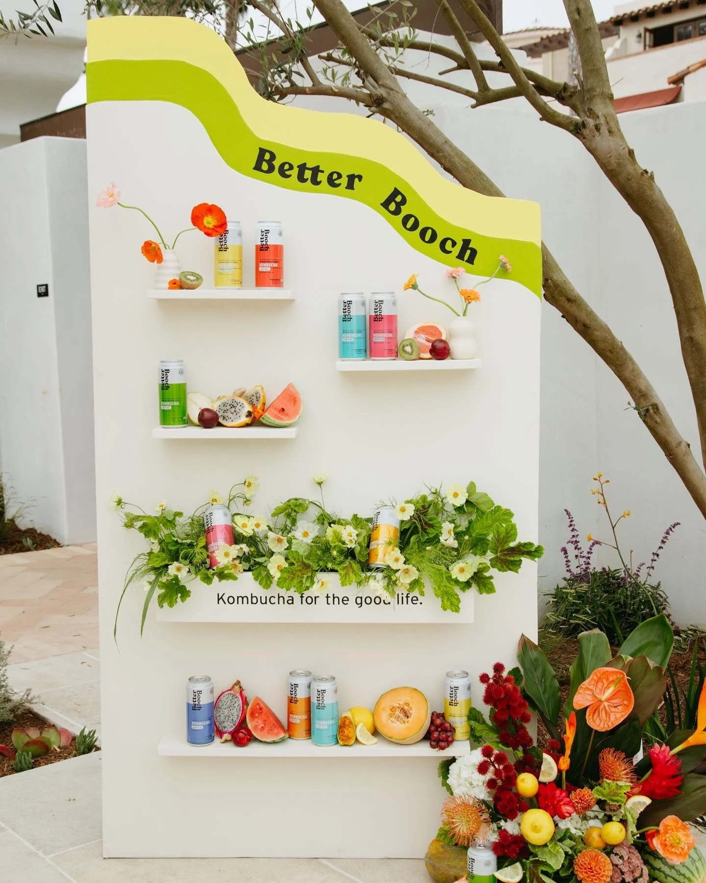 A display of canned beverages, fresh fruits, and flowers arranged on white shelves and a box with the slogan 'Kombucha for the good life.' The display includes cans of kombucha, sliced fruits like watermelon, kiwi, and melon, as well as flowers and g