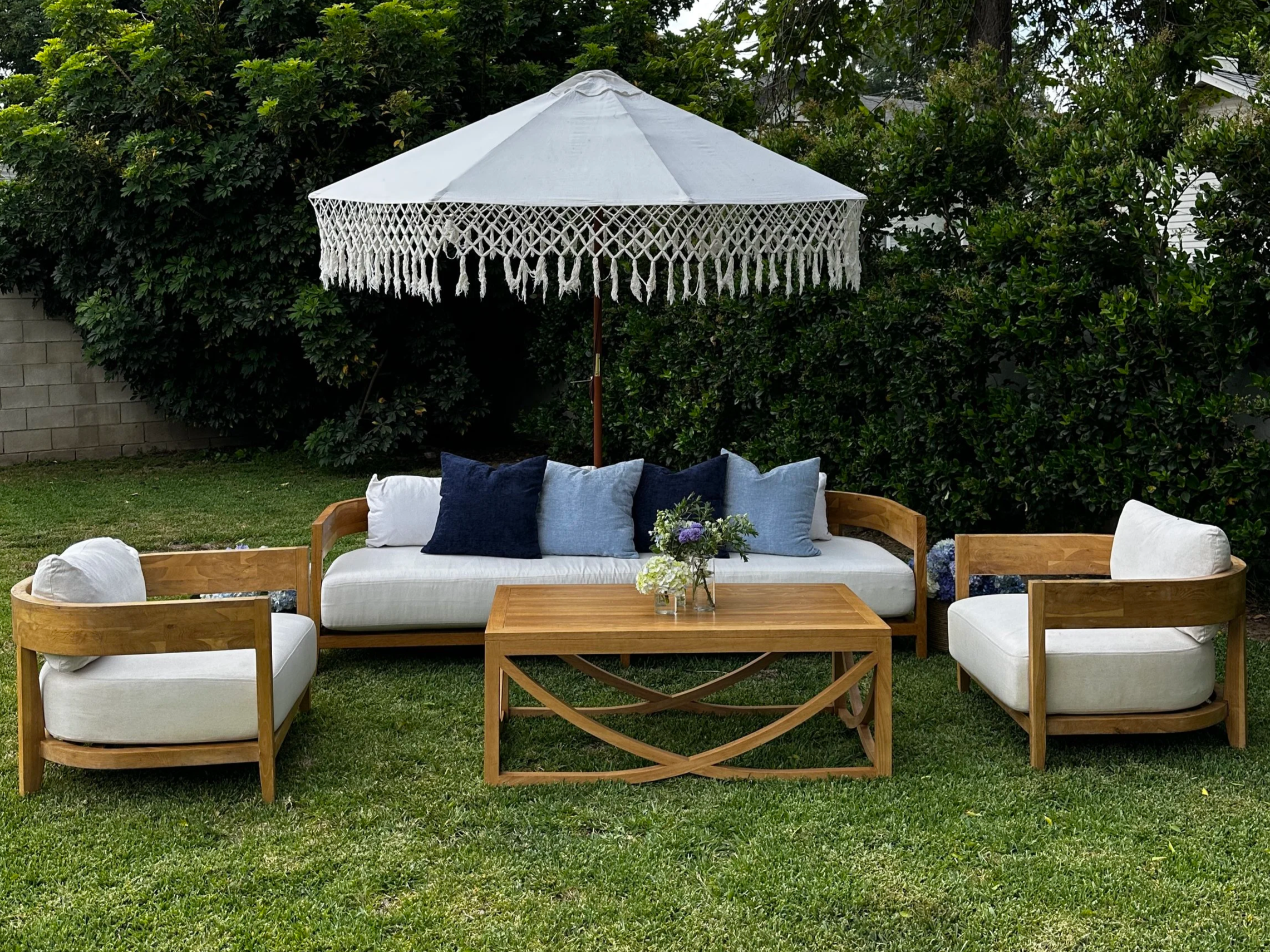 Outdoor seating area with wooden furniture, white cushions, and a white patio umbrella with fringes, set in a grassy backyard with green bushes and trees in the background.