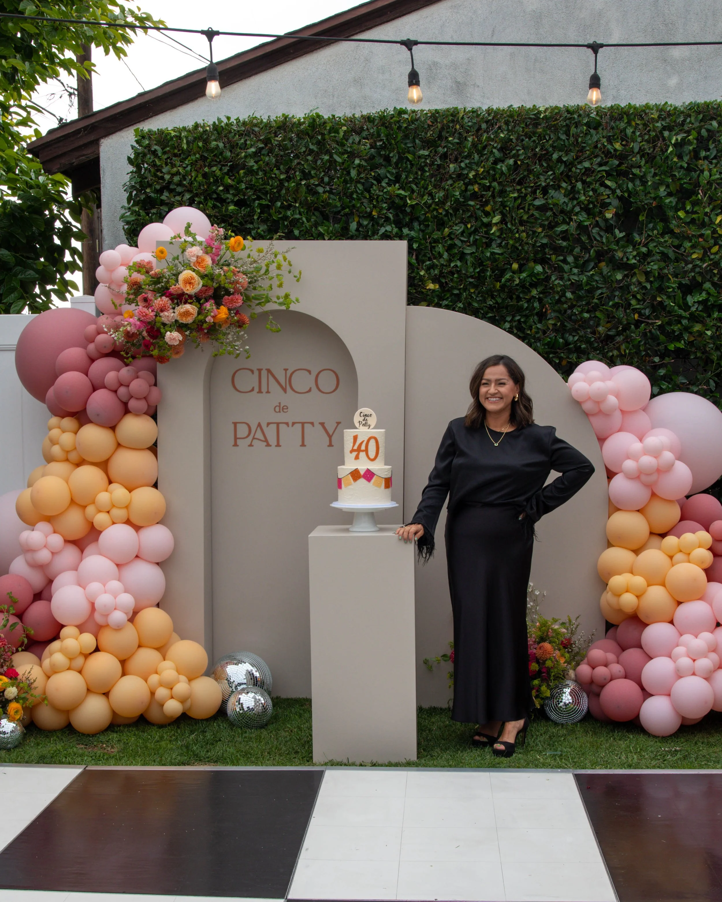 A woman in a black dress stands beside a dessert table with a cake decorated with the number 40, at an outdoor celebration event with pink and peach balloon arch decorations and floral arrangements.