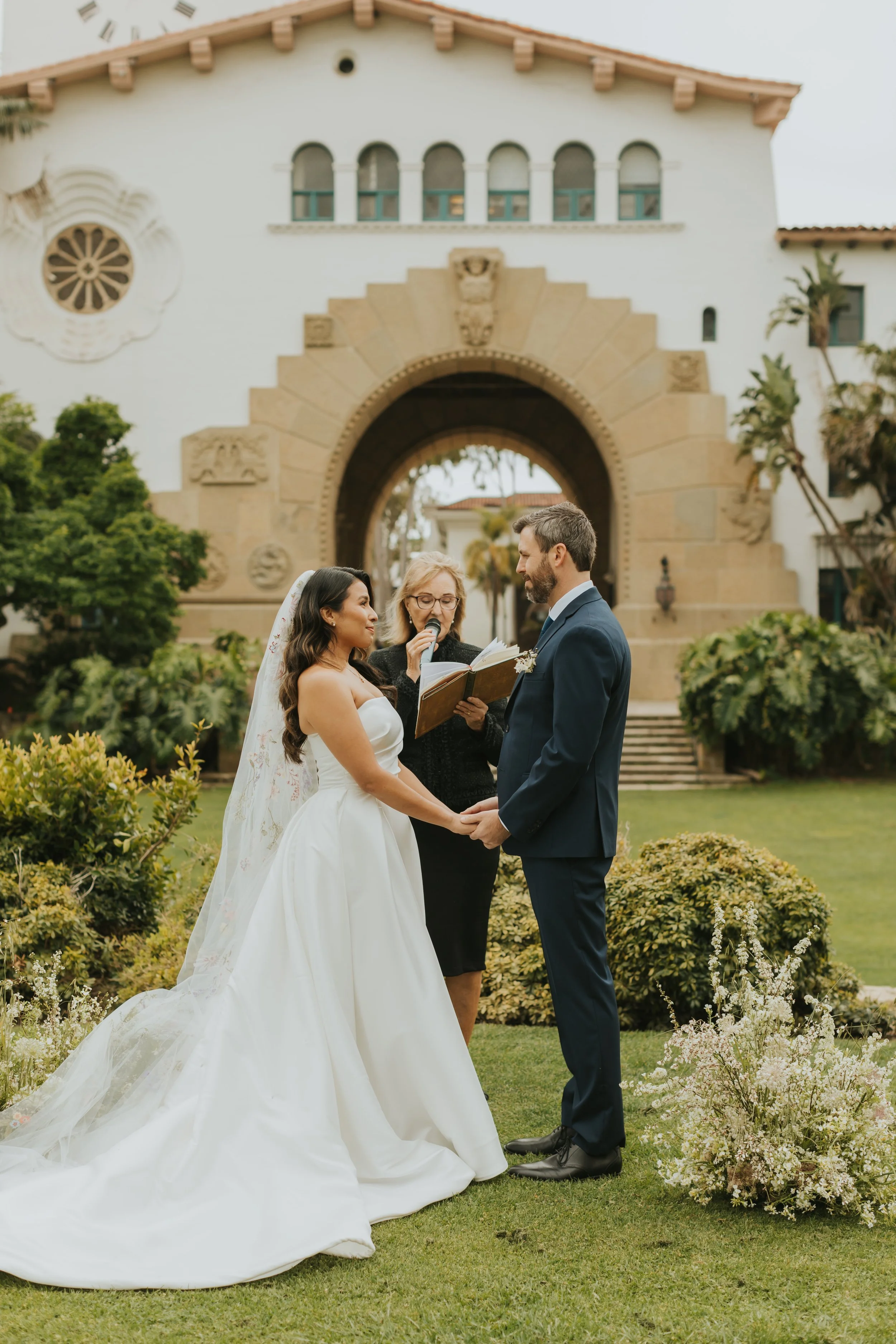 A bride and groom holding hands, standing in front of a church archway during a wedding ceremony, with an officiant reading from a book. The bride is wearing a strapless white wedding gown with a long train, and the groom is in a navy suit. There are