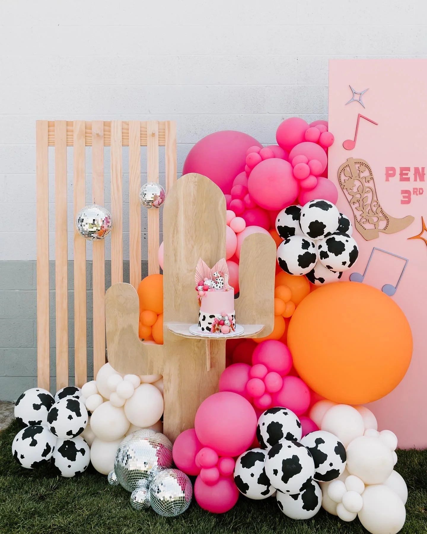 Decorative photo backdrop with pink and white balloons, black and white cow-patterned balloons, and disco balls. There is a wooden panel, a pink birthday cake with a cow pattern and pink decorations, and a pink board with music notes and 'PEN 3RD' wr