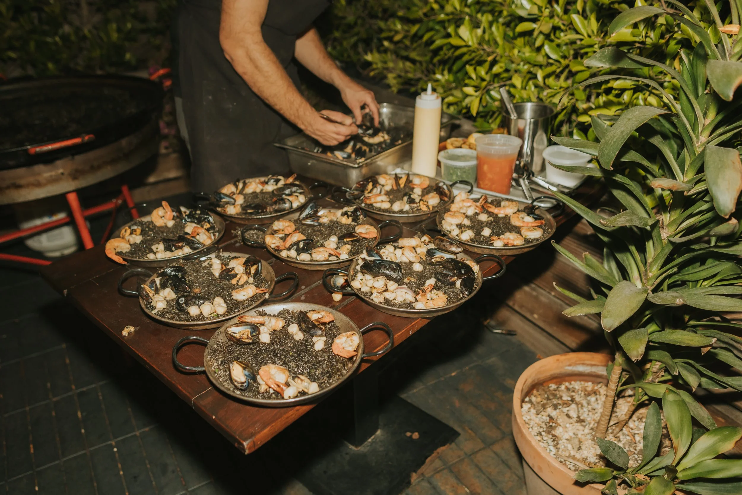 Person preparing shrimp and seafood on outdoor grill at night with potted plants nearby.