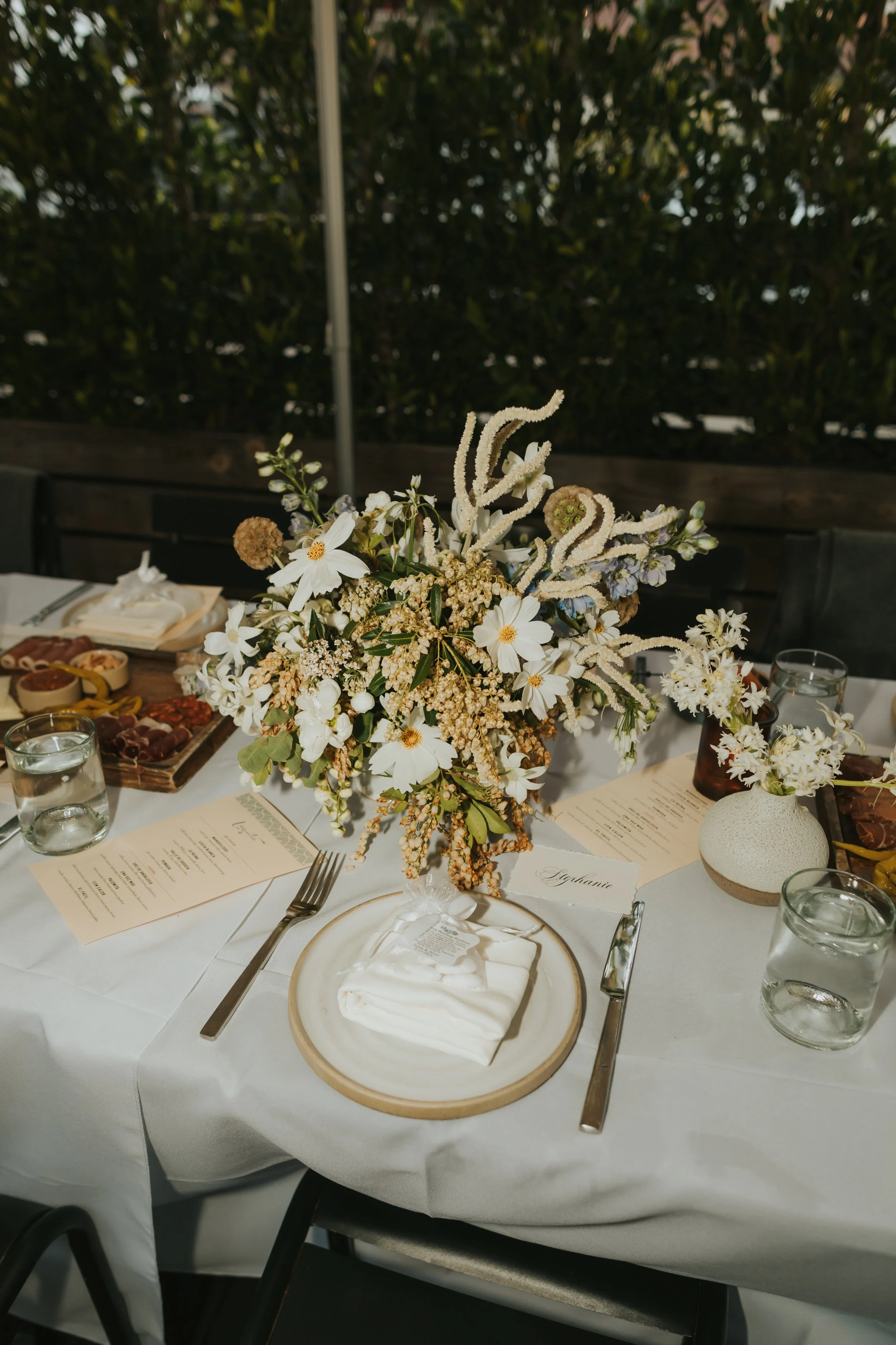 A table setting with a floral centerpiece, place setting with a white napkin wrapped in a ribbon, menu card, glass of water, utensils, and a small ceramic vase, on a white tablecloth, at an outdoor event with a background of greenery.