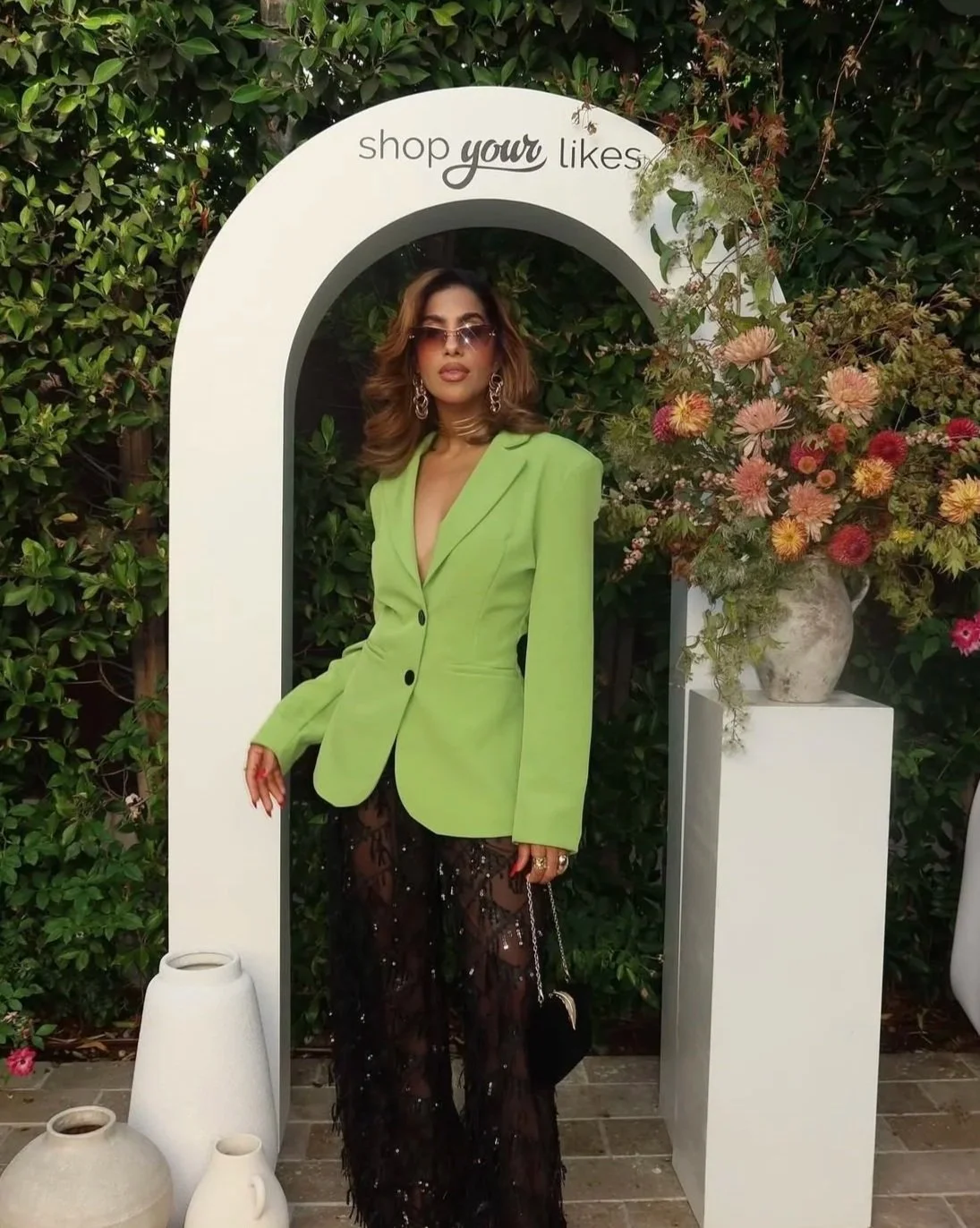 Influencer content creator  in a lime green blazer and black lace pants standing in front of a floral arrangement and greenery at an event with a sign that says "shop your likes."