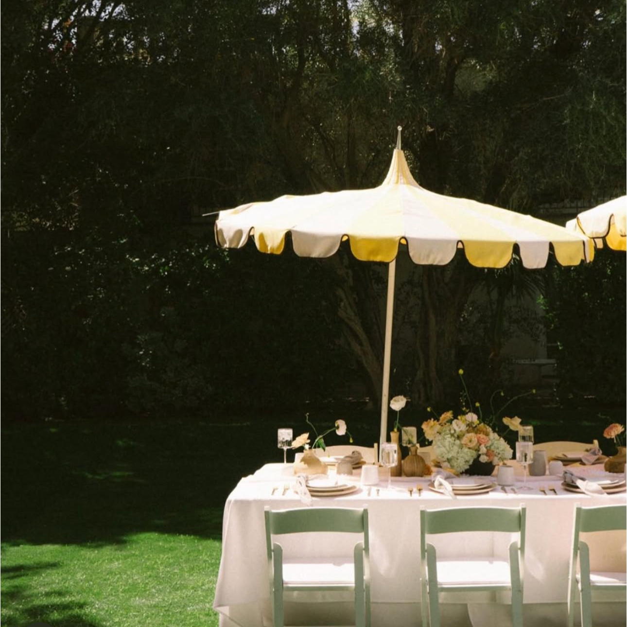 Elegant outdoor dining table under a yellow and white striped umbrella with floral centerpiece, table settings, and chairs on a grassy lawn at night.