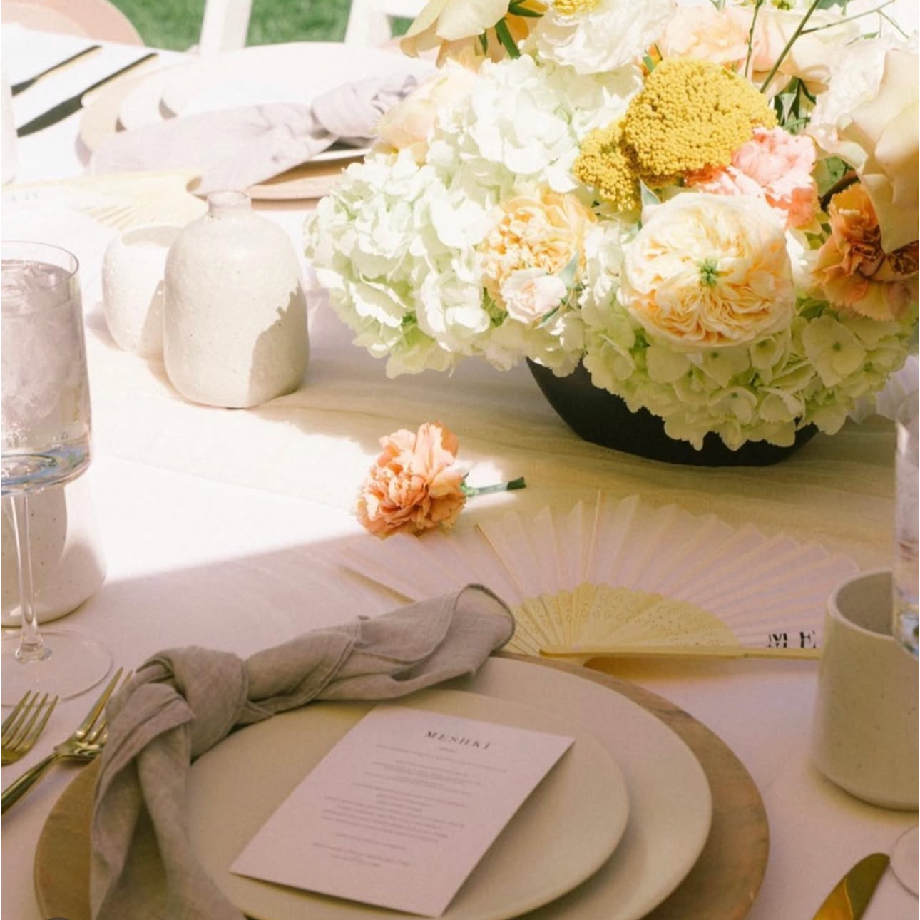 A table decorated with a large bouquet of white and light pink flowers, a white napkin knotted around a fork, a menu card, and various glasses and small decorative items in a bright, outdoor setting.