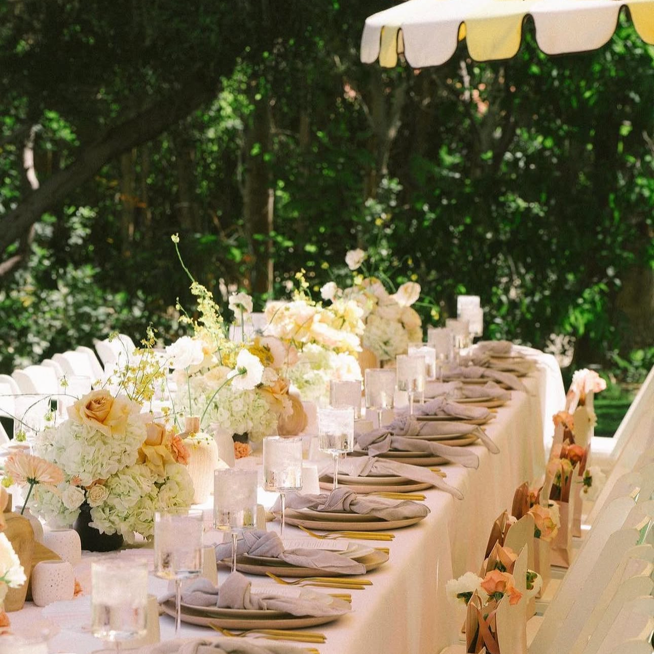 A long outdoor wedding reception table decorated with white and blush floral arrangements, candles, and place settings, set against a lush green background.