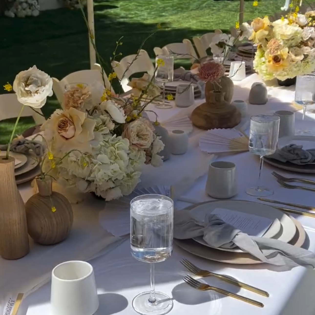 A beautifully arranged outdoor dining table decorated with white and blush floral centerpieces, wooden vases, candles, white tableware, and gold utensils, set in a garden setting.