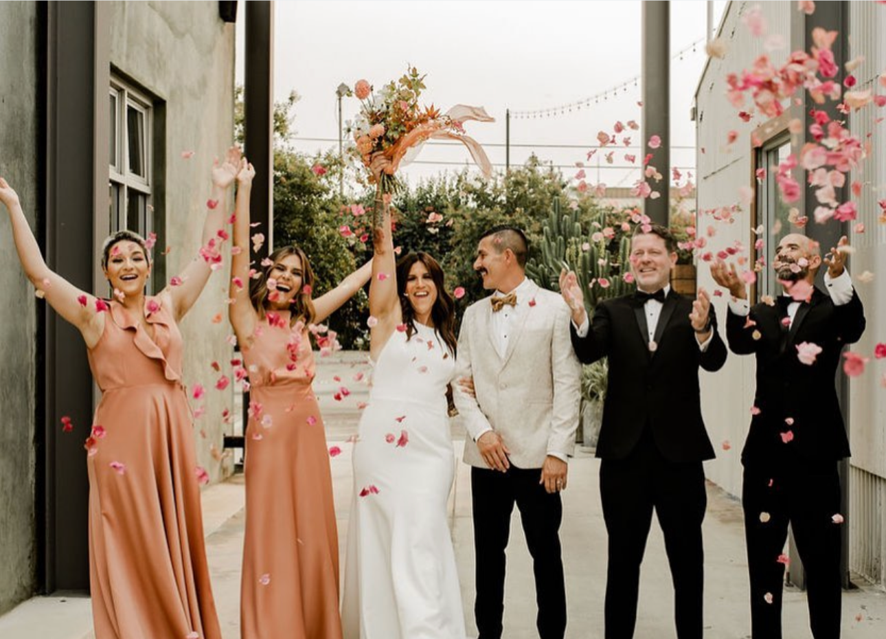 Bride and groom celebrating with wedding party as rose petals fall in an outdoor alleyway.