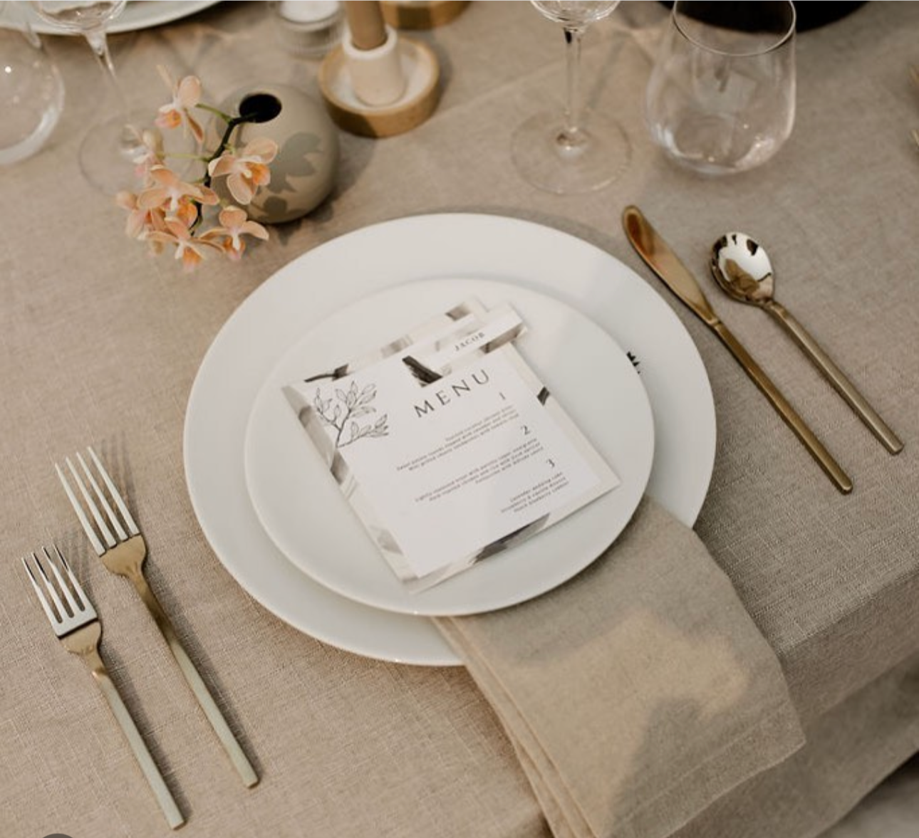 Elegant table setting with a white plate, stainless steel cutlery, a menu, and decorative items including flowers in a vase, candles, and wine glasses.
