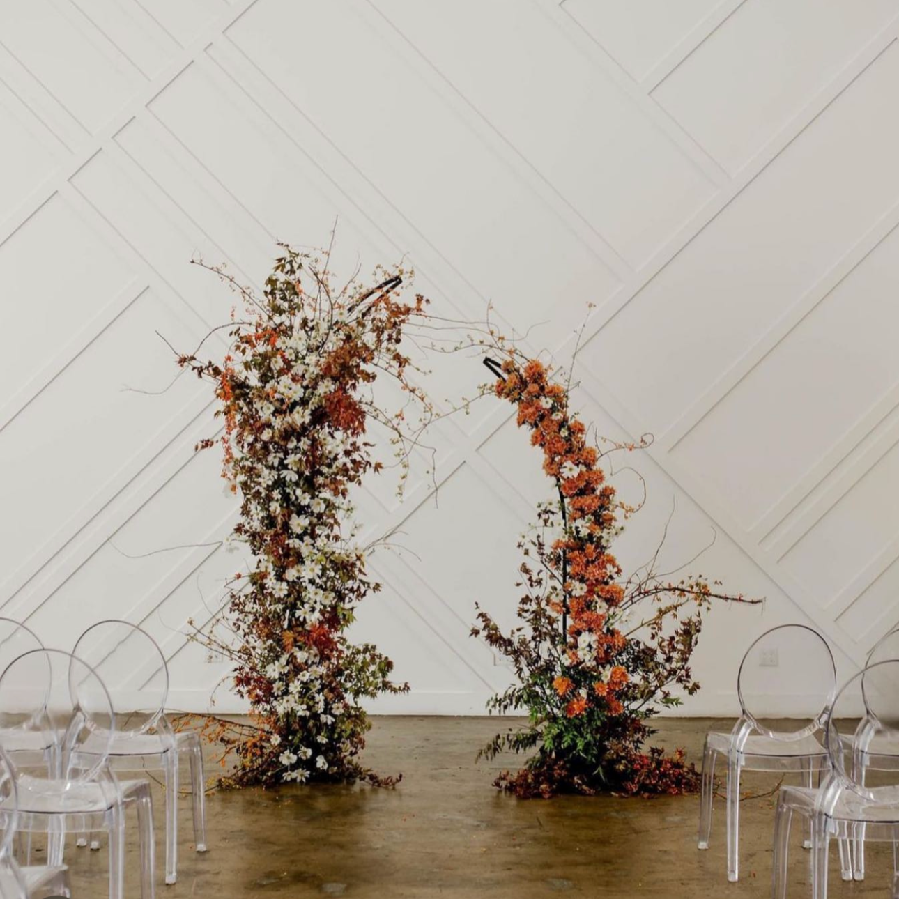 A floral arch with autumn leaves and white and orange flowers, set against a white wall, with transparent chairs nearby.