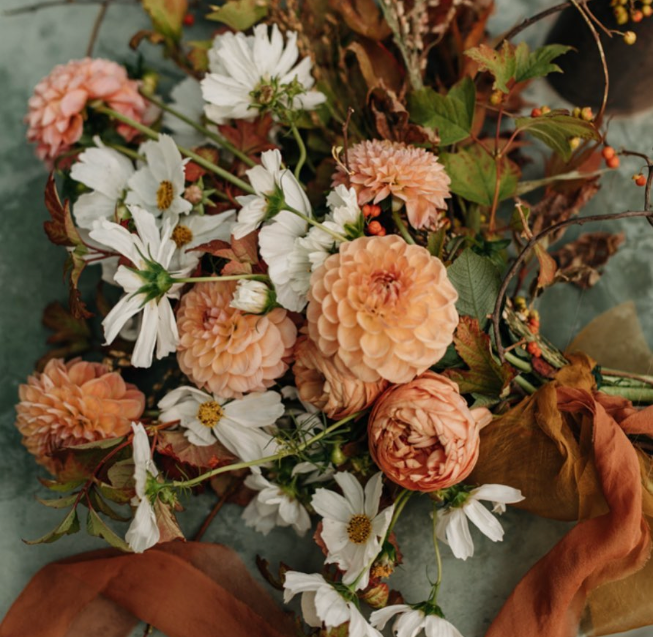 A bouquet of peach-colored dahlias and white daisies with green leaves and brown dried foliage on a textured surface.