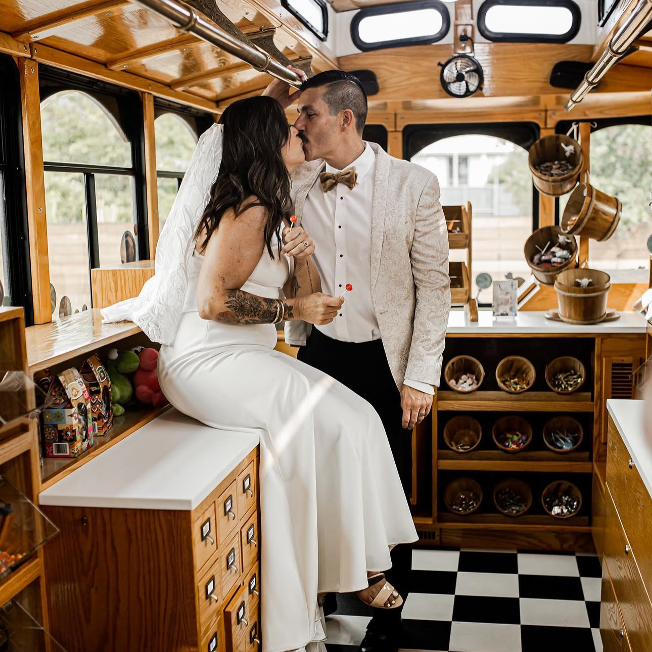A bride and groom sharing a kiss inside a vintage wooden trolley, with shelves of candy and snacks around them.