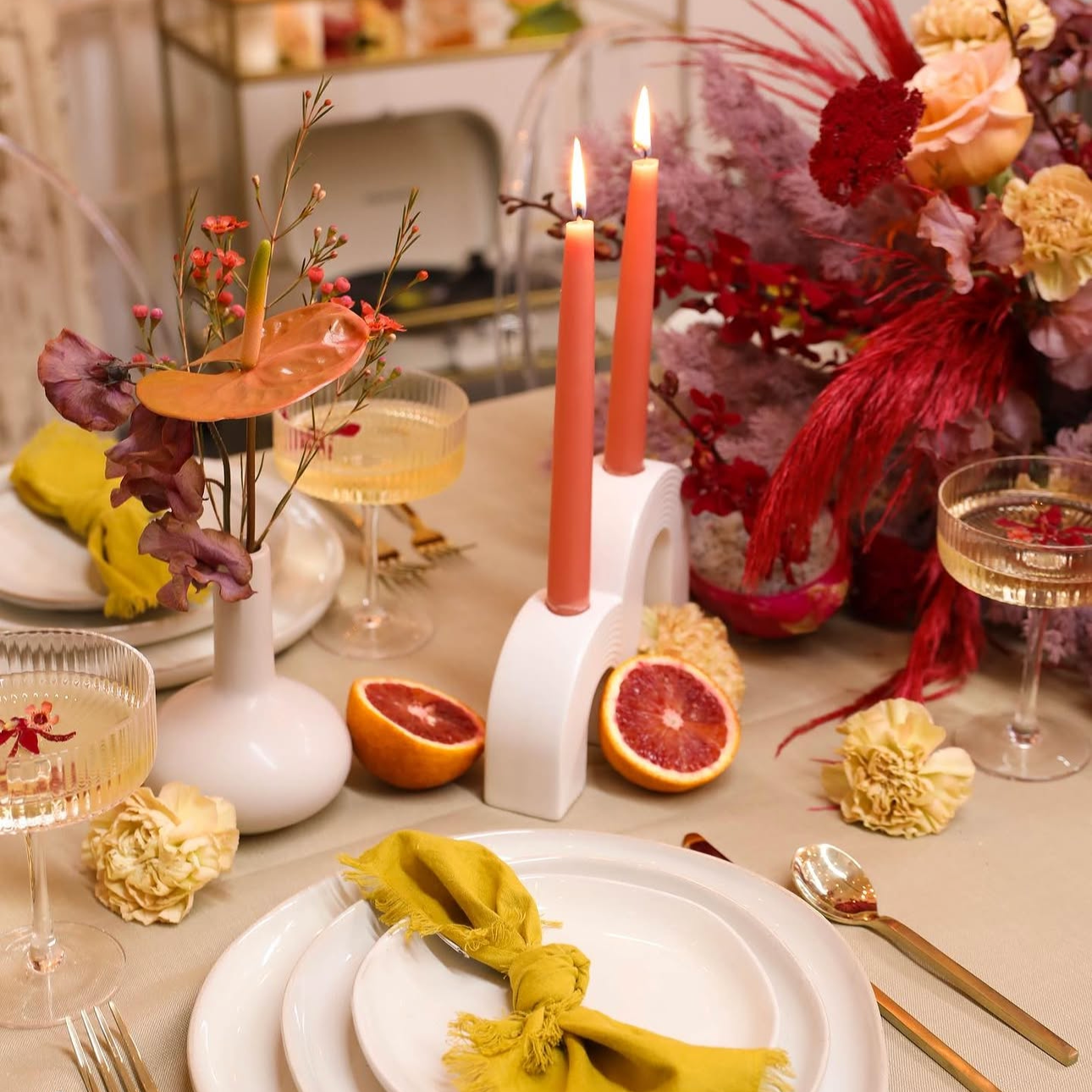 A decorated dining table with pink candles, floral arrangements, halved grapefruits, and vintage-style glasses. The table is set with white plates and gold flatware, and a yellow cloth napkin tied into a knot.