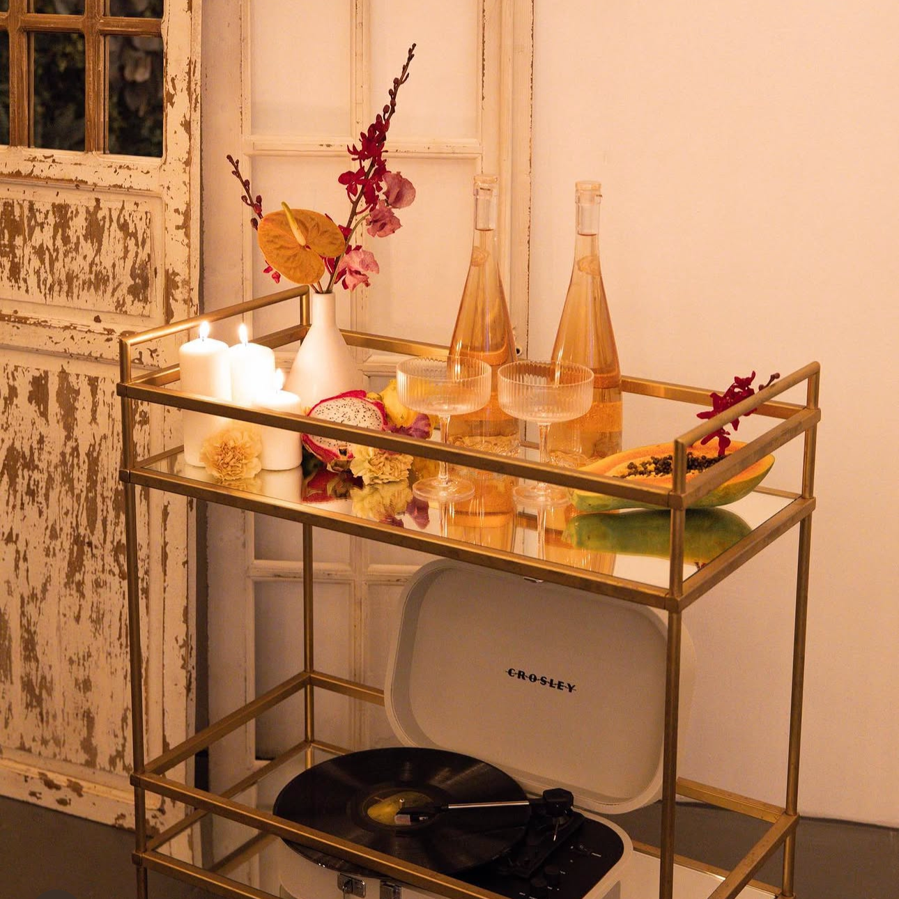 A gold-framed bar cart with decorative items including candles, flowers in a white vase, two glass bottles, a couple of cocktail glasses, a sliced dragon fruit, and a papaya with black seeds, set against a distressed wooden door and a plain wall.
