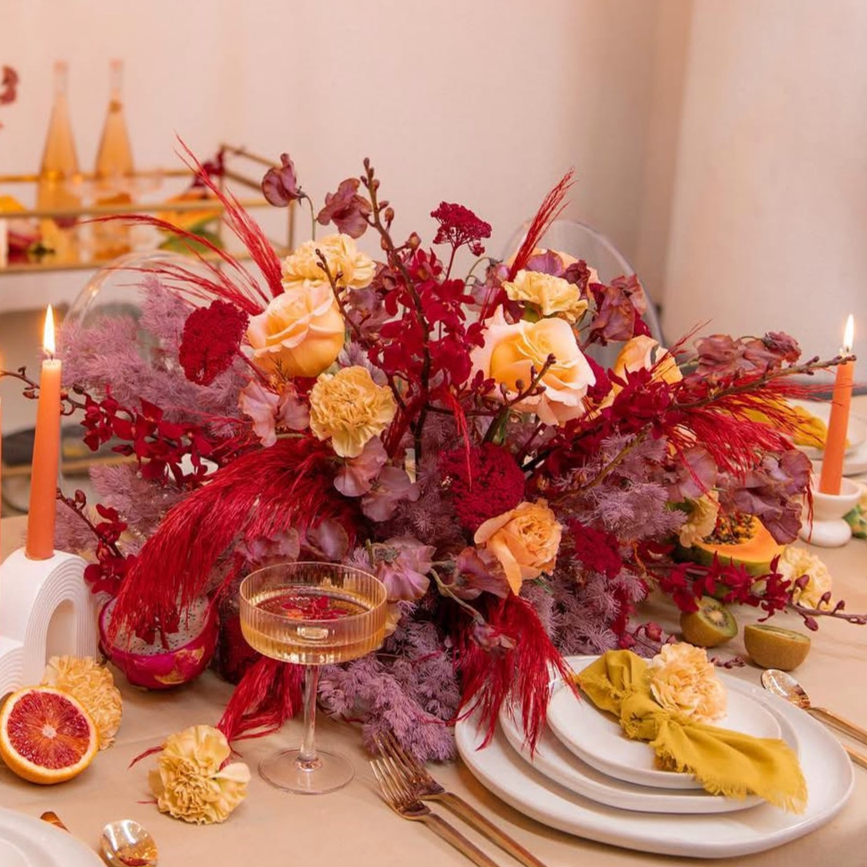 A table centerpiece of red, pink, and peach flowers with candles and place settings.
