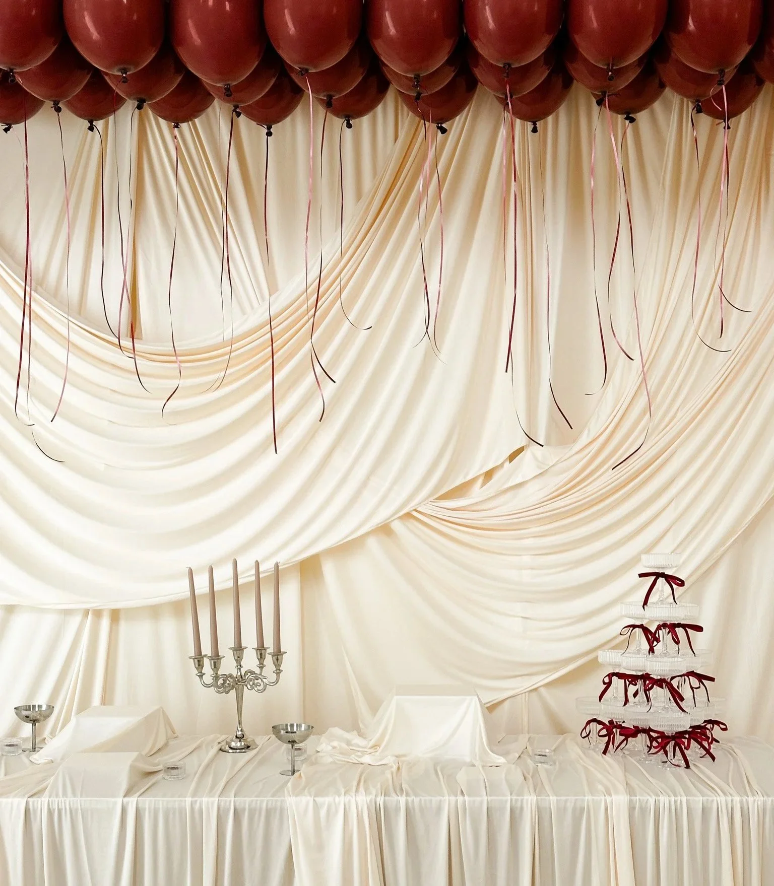 Elegant banquet table setup with a cream-colored draped backdrop, a candelabra with five tall candles, tiered display trays with wrapped treats decorated with red ribbons, and a white tablecloth.