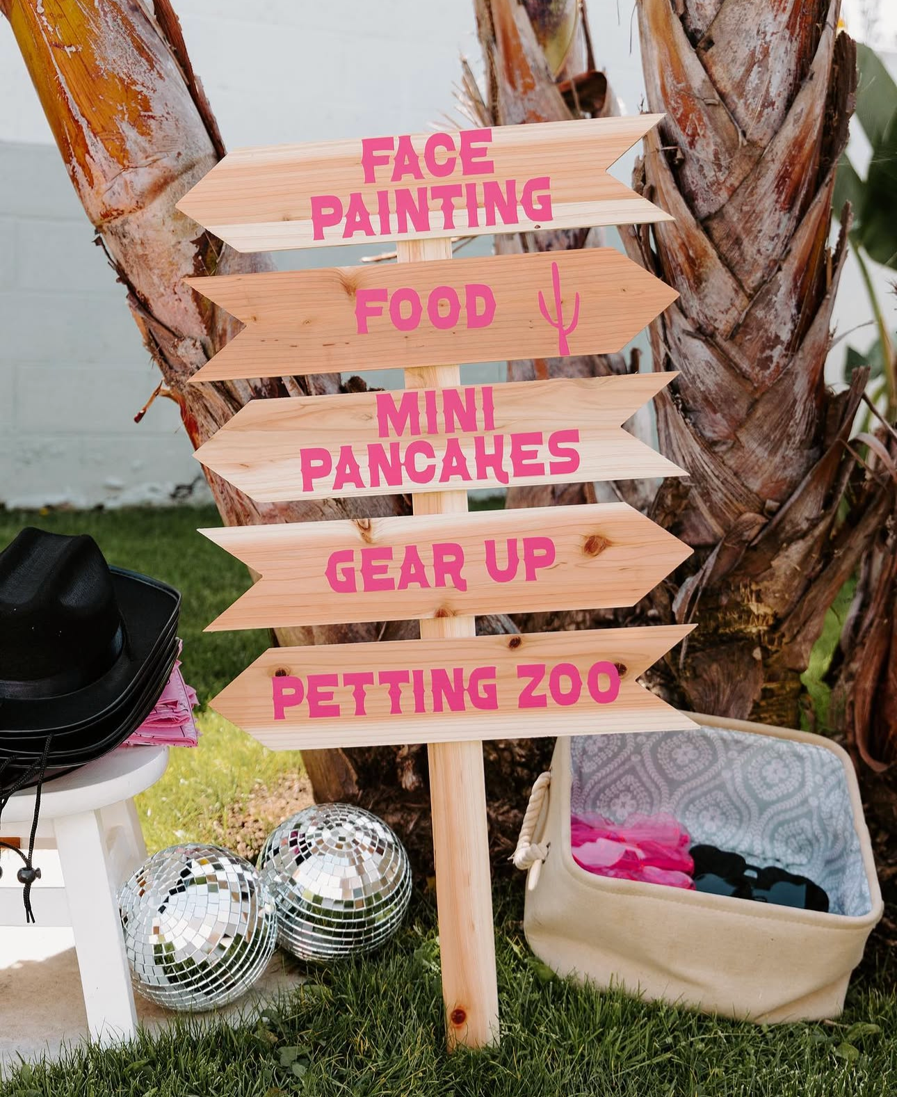 Wooden directional signs with pink lettering and arrows pointing to various activities such as face painting, food, mini pancakes, gear up, petting zoo; signs are placed outdoors near a palm tree, with disco balls, a white stool with a black hat, and