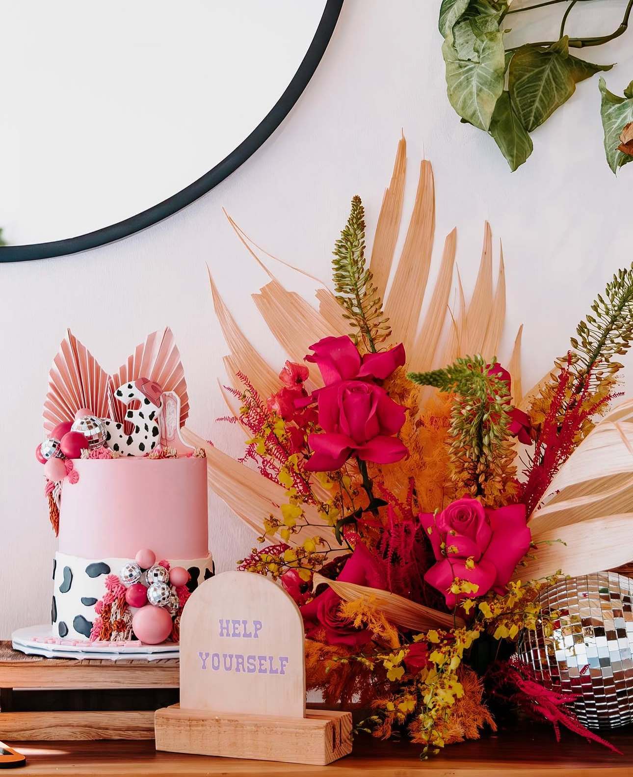Decorative cake with pink, black, and white color scheme, topped with a cow print number one, pink and silver balls, and pink flowers, set on a wooden table. Behind it is a large pink and leopard print wedding cake with pink accents. To the right, a 