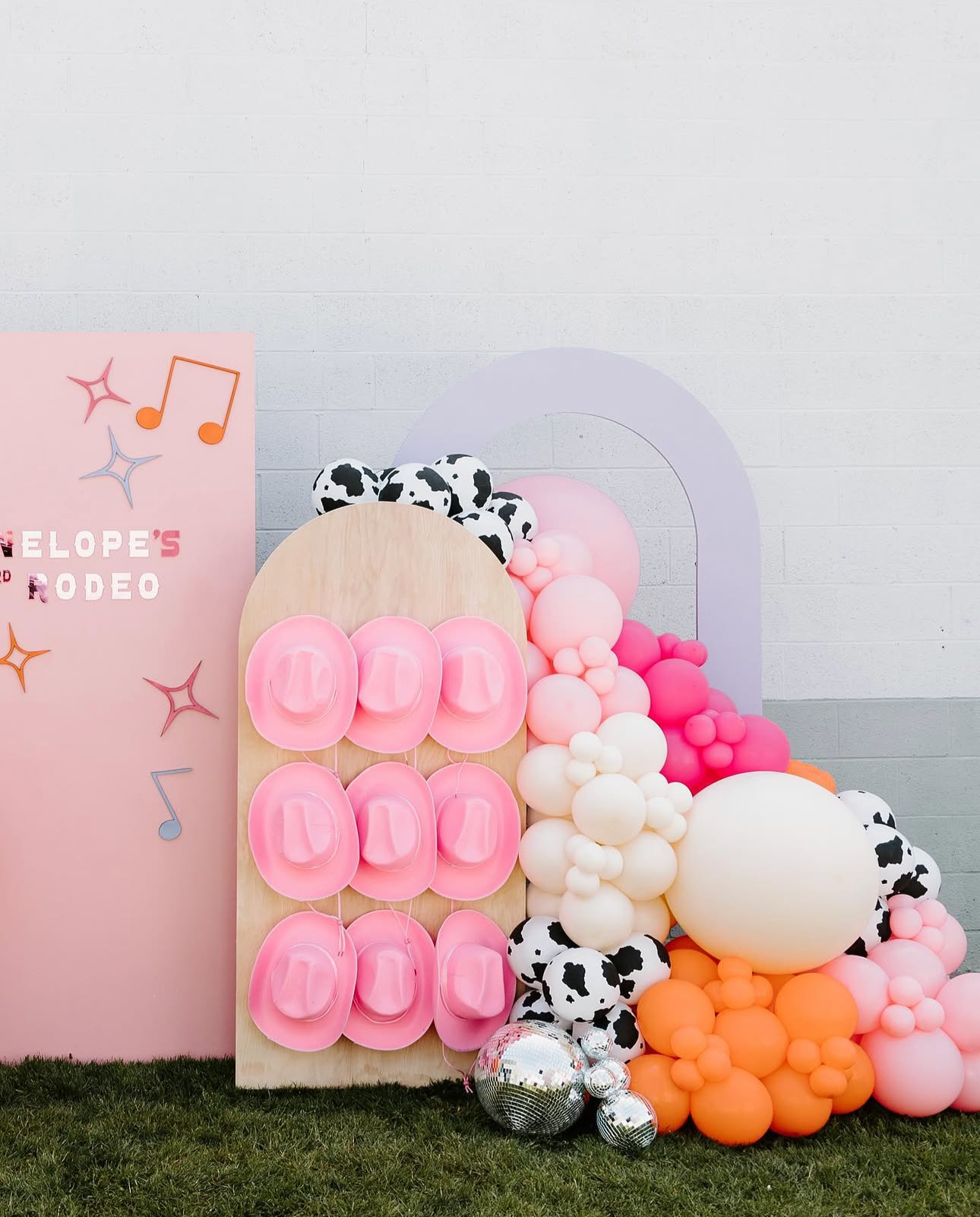 Pink and orange balloon arrangement with a wooden panel displaying pink plastic shoes, set against a pastel pink backdrop with musical notes and star decorations.