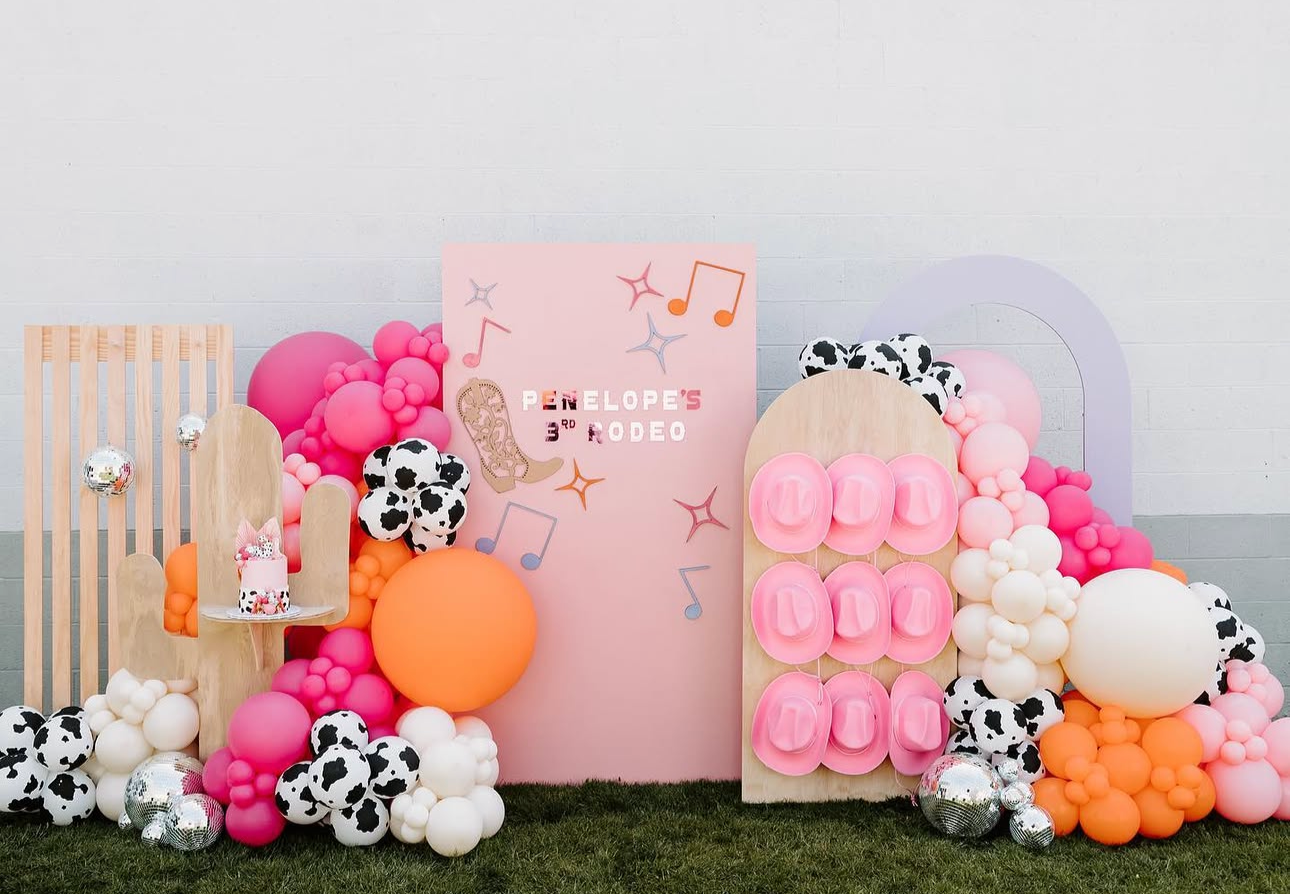 Colorful party decoration with pink, orange, white, and cow print balloons, wooden and cardboard props, and a pink sign that reads "Penelope's 3rd Rodeo" with musical notes and cowboy boot illustrations.
