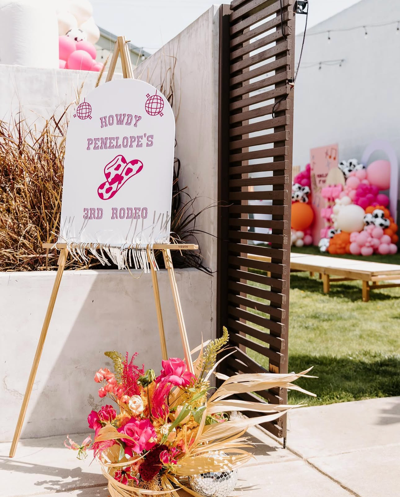 A white sign with pink text and images reading "Howdy Penelope's 3rd Rodeo" is displayed on an easel. A colorful floral arrangement with pink, red, blonde, and orange flowers is in front of the sign. In the background, there are pink and black-and-wh