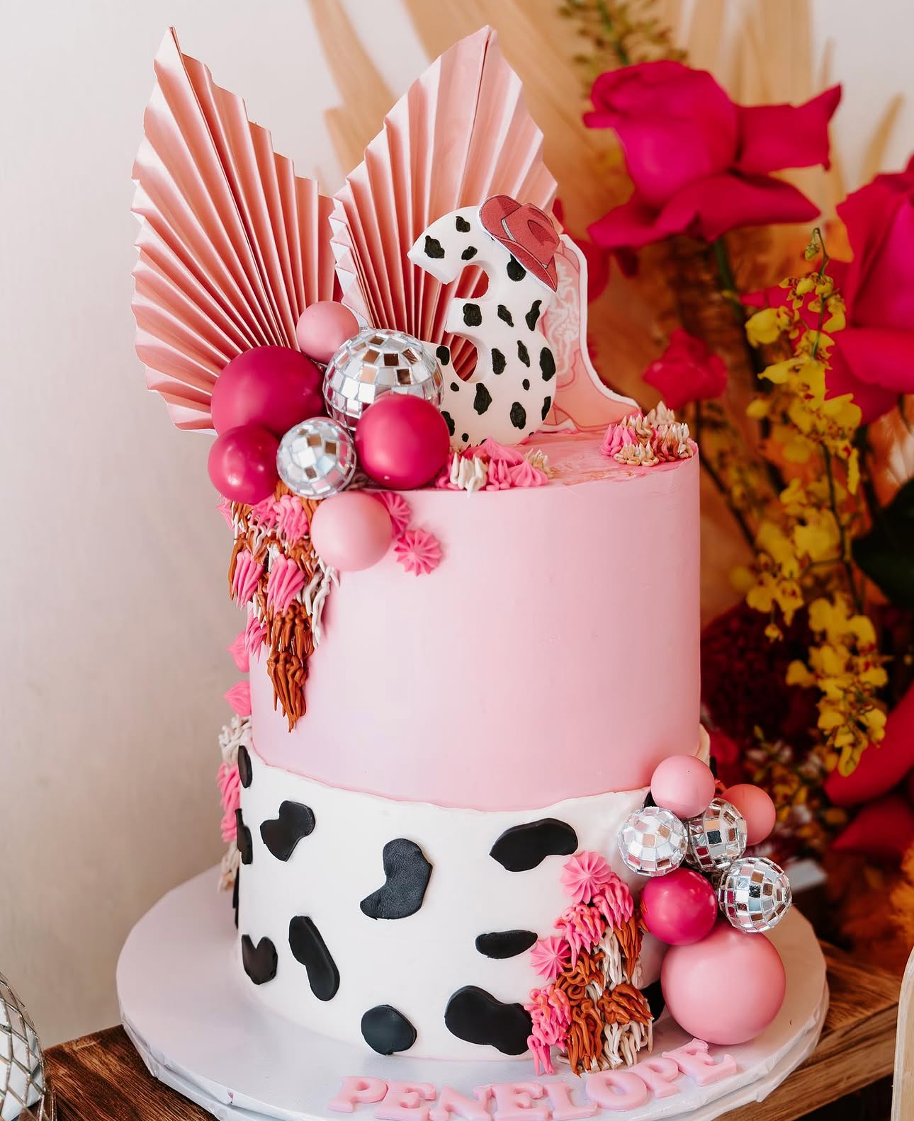 Pink and white tiered cake decorated with cow print, pink buttercream flowers, pink and silver balls, a cow figurine with a red hat, and large pink paper fans, with pink flowers and more decorations in the background.