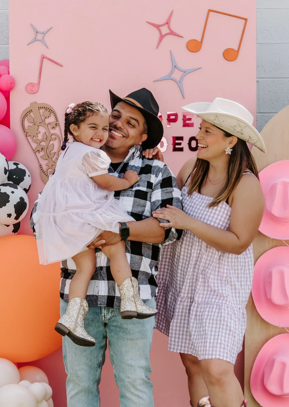 A family celebrating a birthday party with a pink background, musical note decorations, balloons, and a cow-themed decoration. The man is holding a young girl, both smiling, while the woman looks on happily.