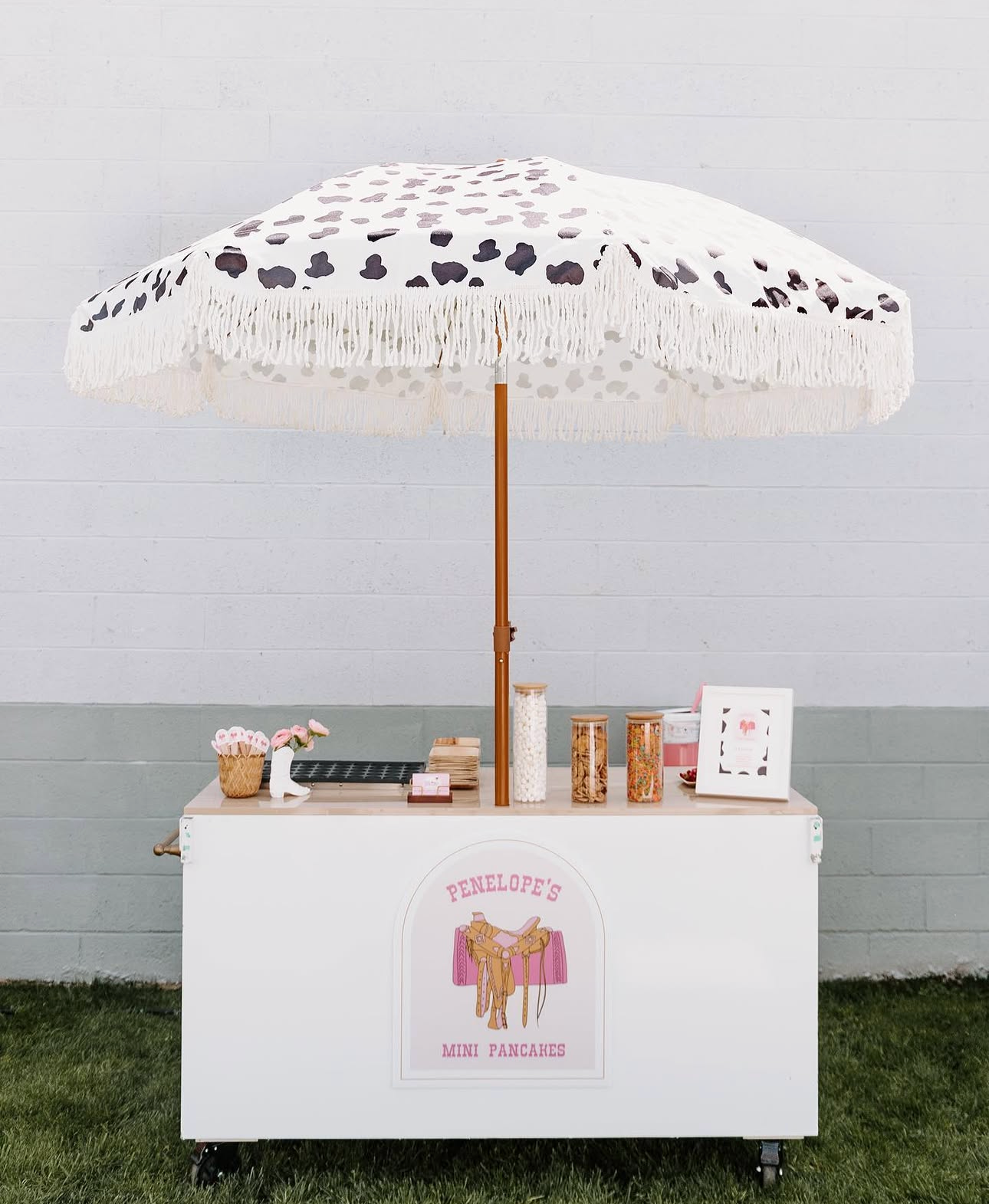 A white cart with a pink sign that reads 'Penelope's Mini Pancakes' and features a graphic of a pink overturned suitcase with clothes. On top of the cart is a large beach umbrella with a white background and black and gray spots, decorated with white