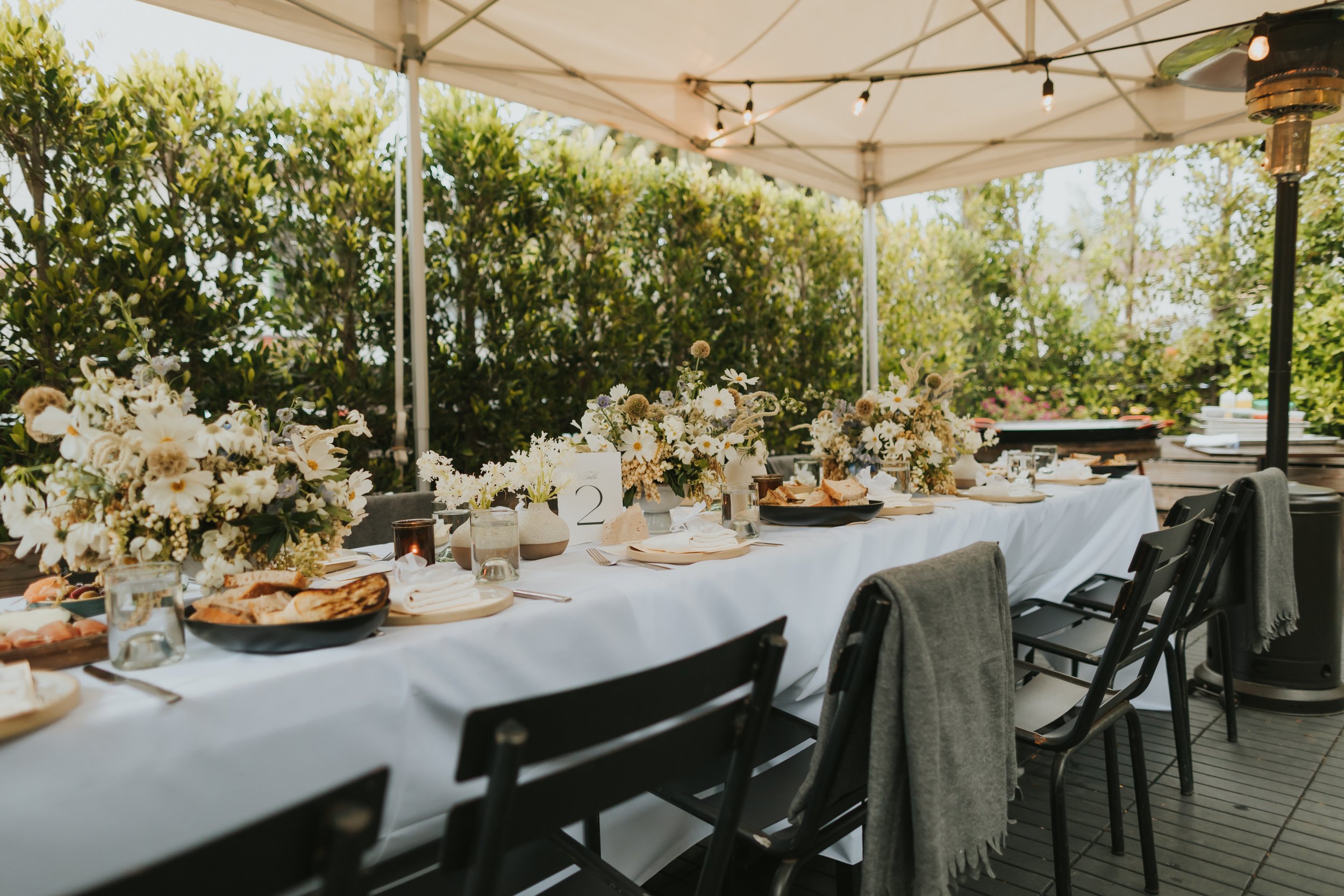 Outdoor dining setup under a white canopy with a long table decorated with white and beige flowers, candles, plates, and food, surrounded by black chairs, with greenery in the background.