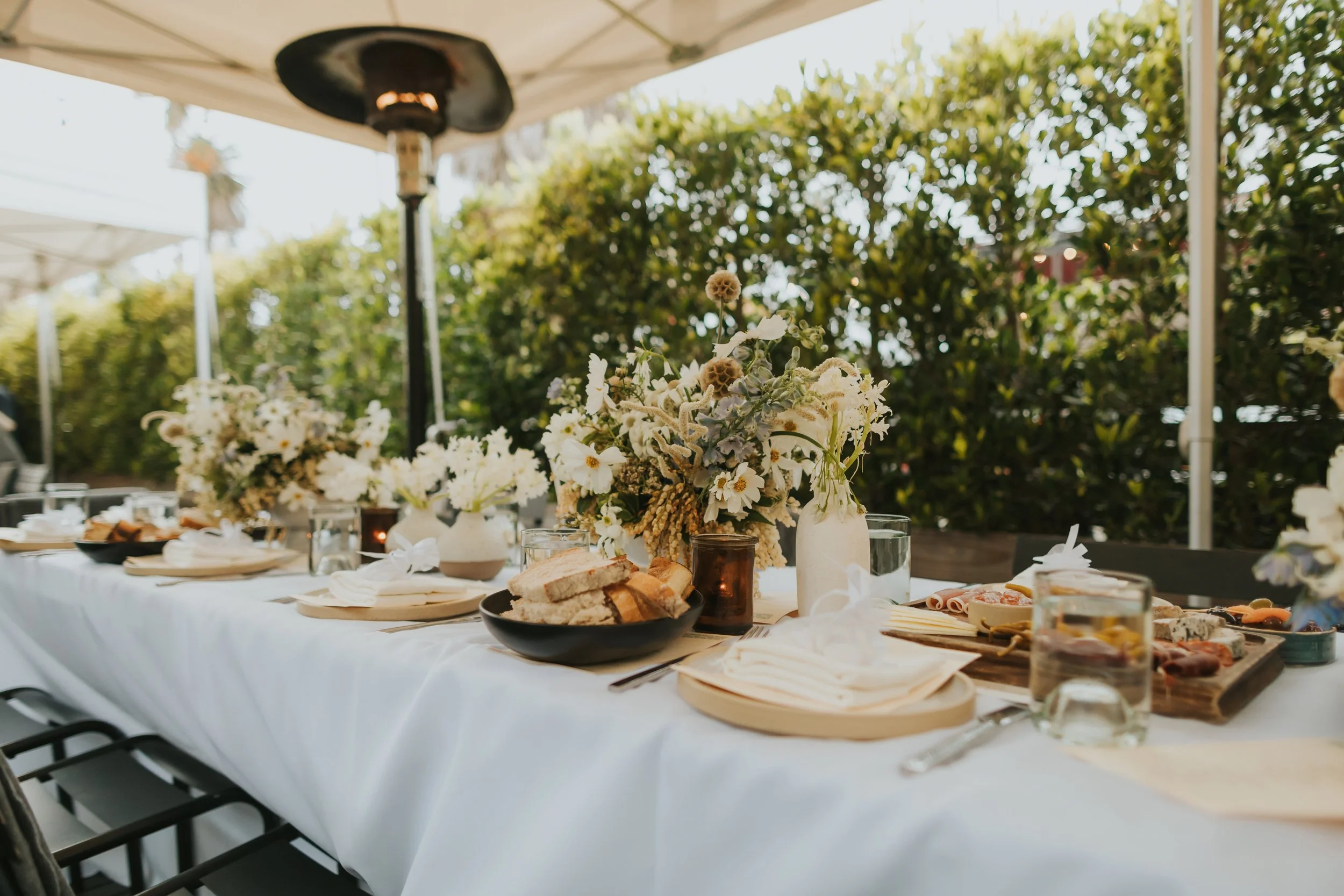 A table set for an outdoor meal with white flowers in vases, bread, and various dishes on a white tablecloth under a tent with greenery in the background.
