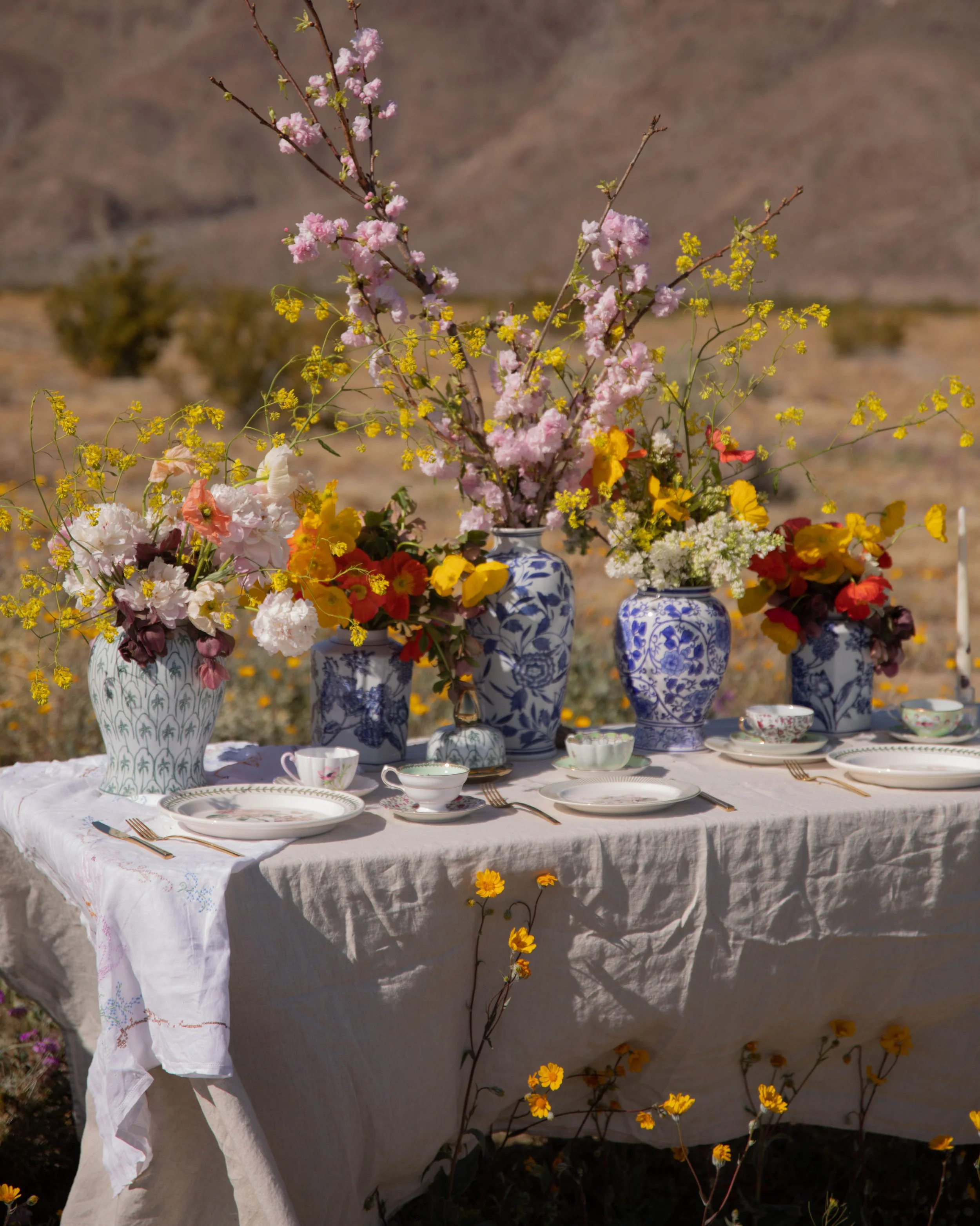 A table with a white tablecloth set for a meal, featuring blue and white patterned vases filled with pink, yellow, orange, and white flowers, in a desert landscape with rocks and sparse vegetation.