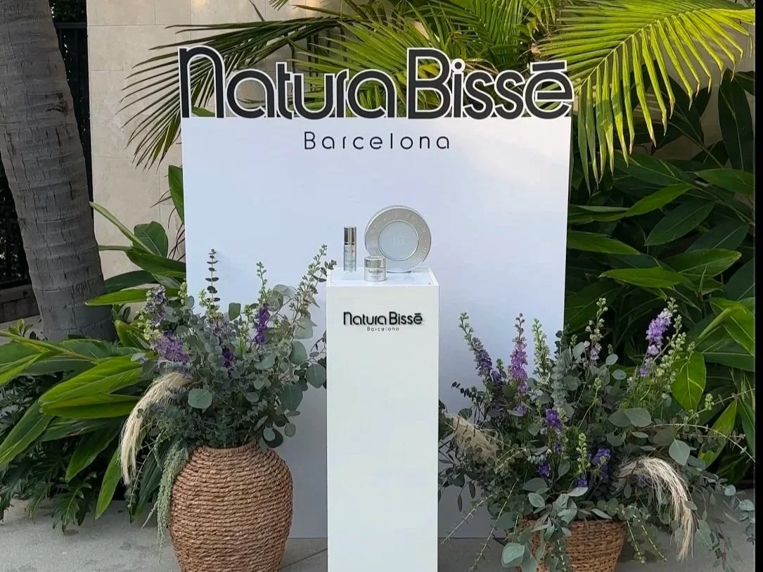 Beauty products displayed on a white pedestal with the sign 'Natura Bissé Barcelona' behind, surrounded by potted purple and green foliage, with a white backdrop and tropical leaves.