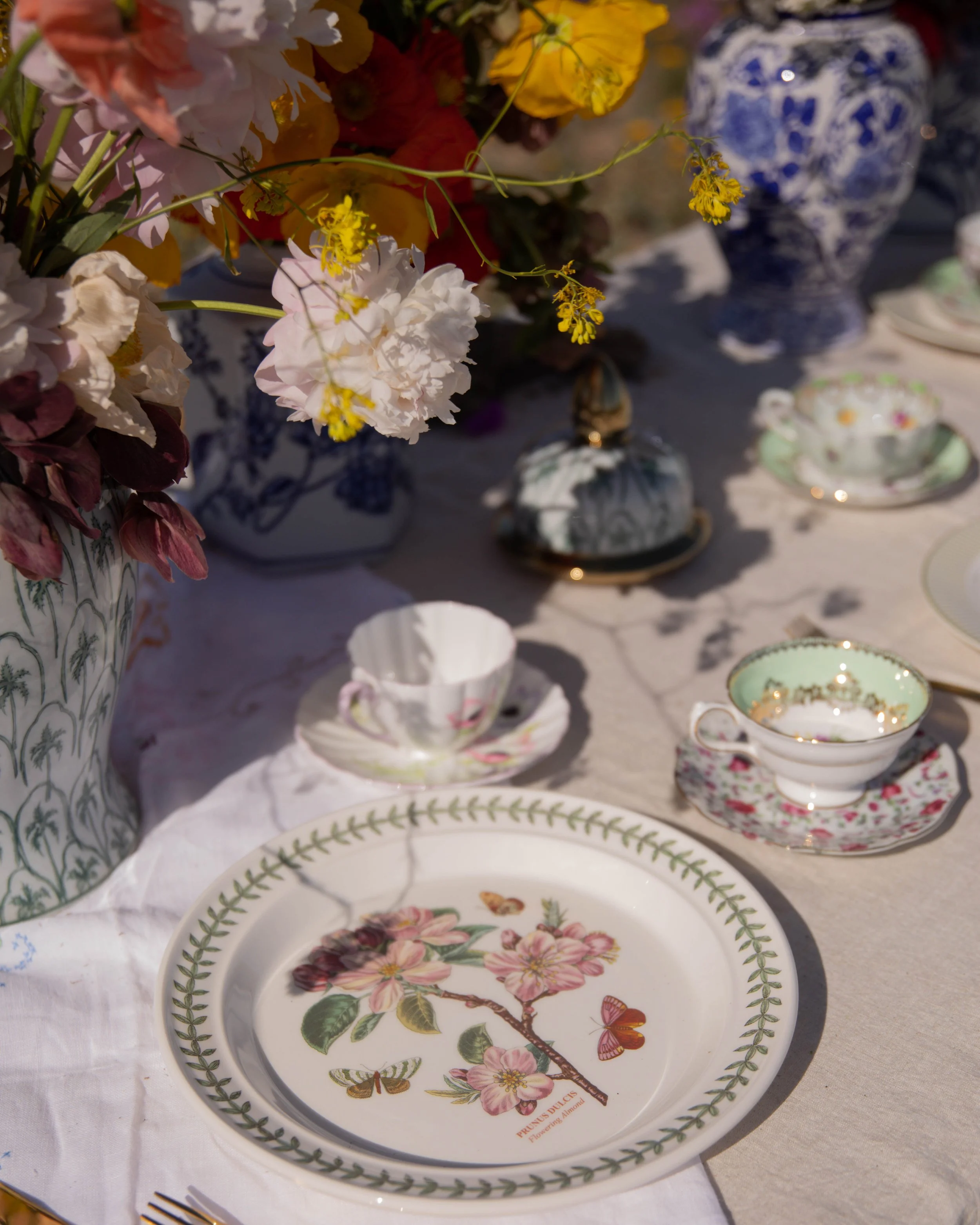 A decorative floral plate featuring cherry blossoms and butterflies, with a green vine border, on a white tablecloth in a tea set arrangement with other fine china in the background.