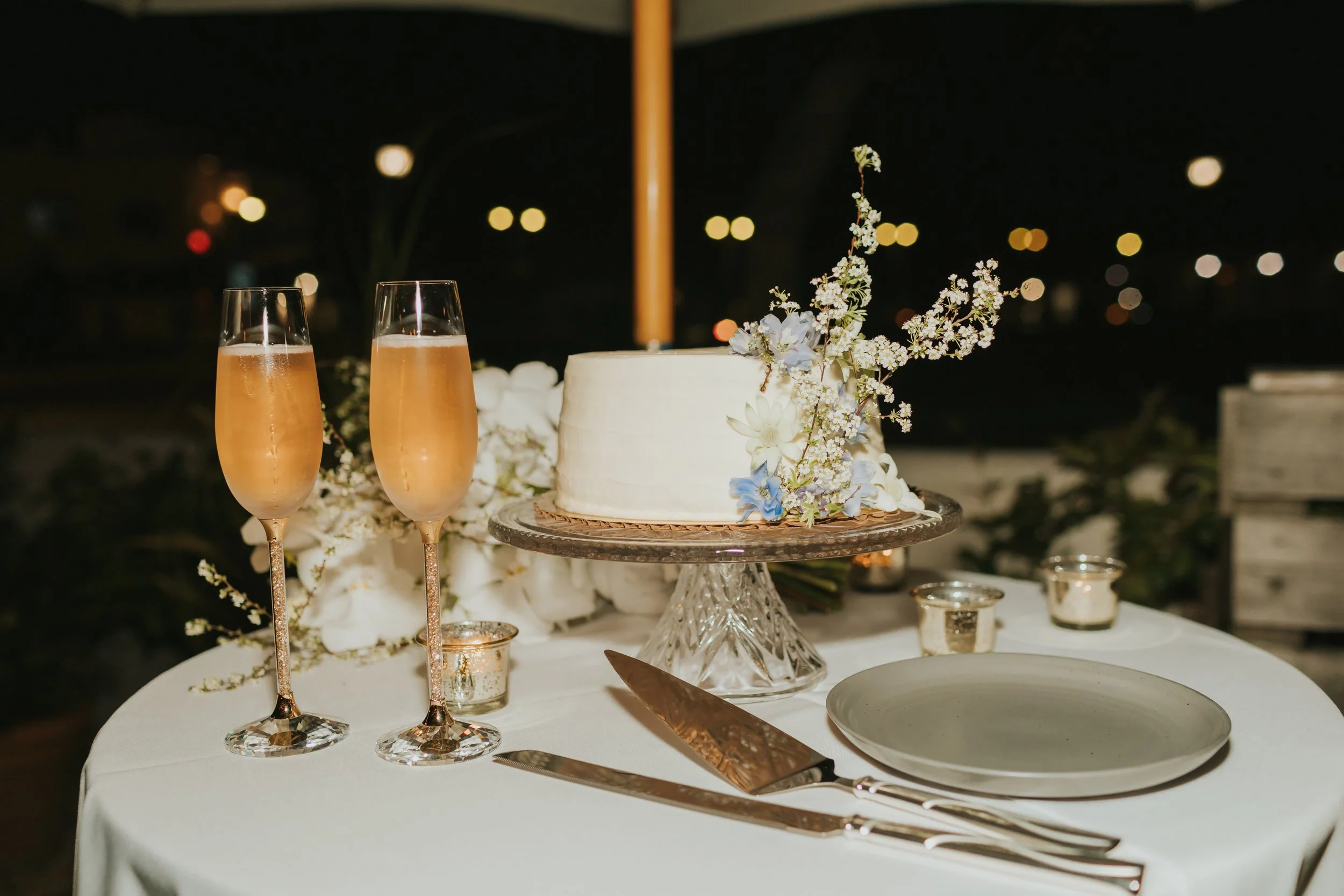 Wedding cake decorated with white and light purple flowers on a glass stand, flanked by two champagne flutes filled with champagne, on a white tablecloth at night with blurred lights in the background.
